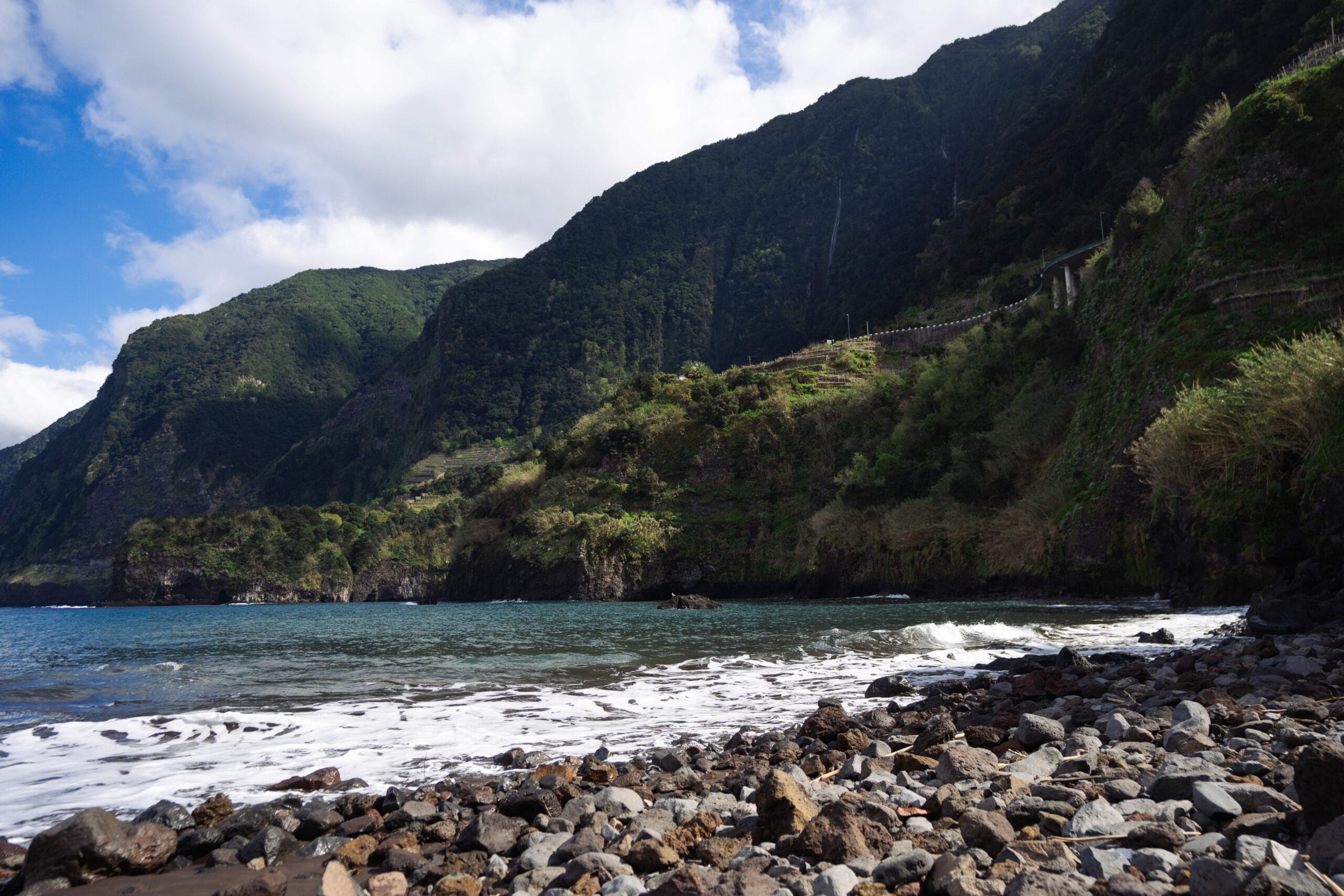 Rocky coastline with gentle waves meets lush, green mountains under a partly cloudy sky, creating a serene and picturesque landscape.