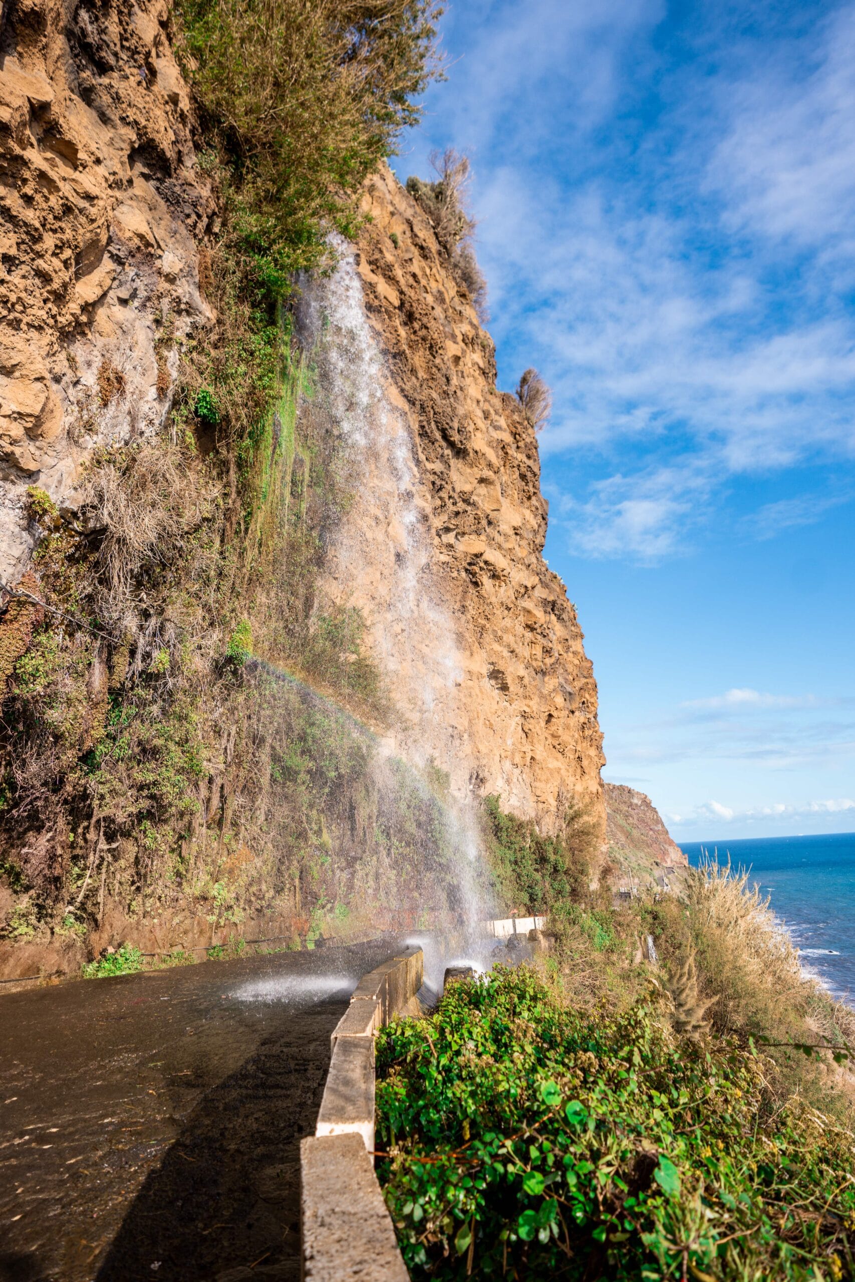 Cliffside waterfall cascading down over rocks with green foliage, creating a small rainbow. Bright blue sky and ocean in the background, conveying tranquility.
