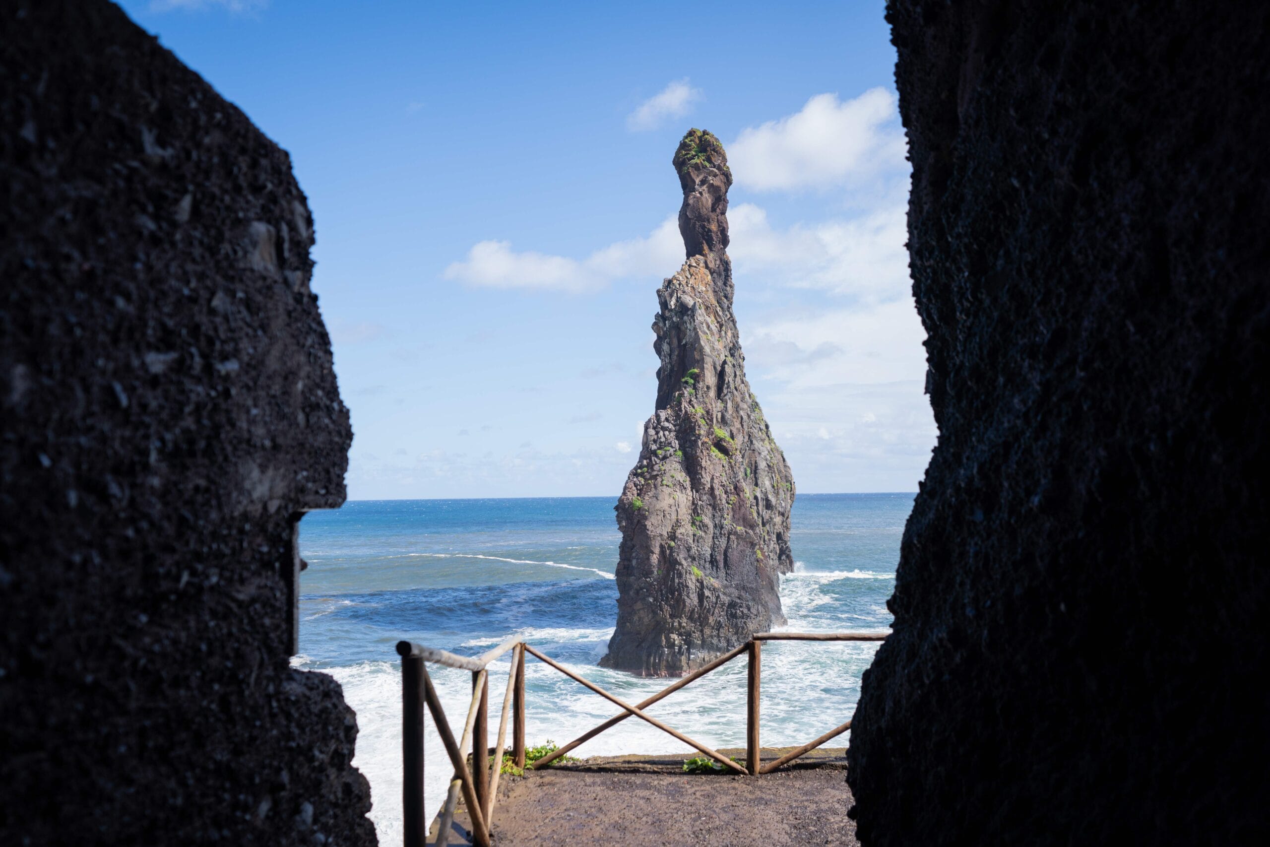 Tall rock formation rises from the ocean, viewed through a coastal cave opening. Rough cliffs frame the view; wooden fence in foreground. Bright, serene scene.