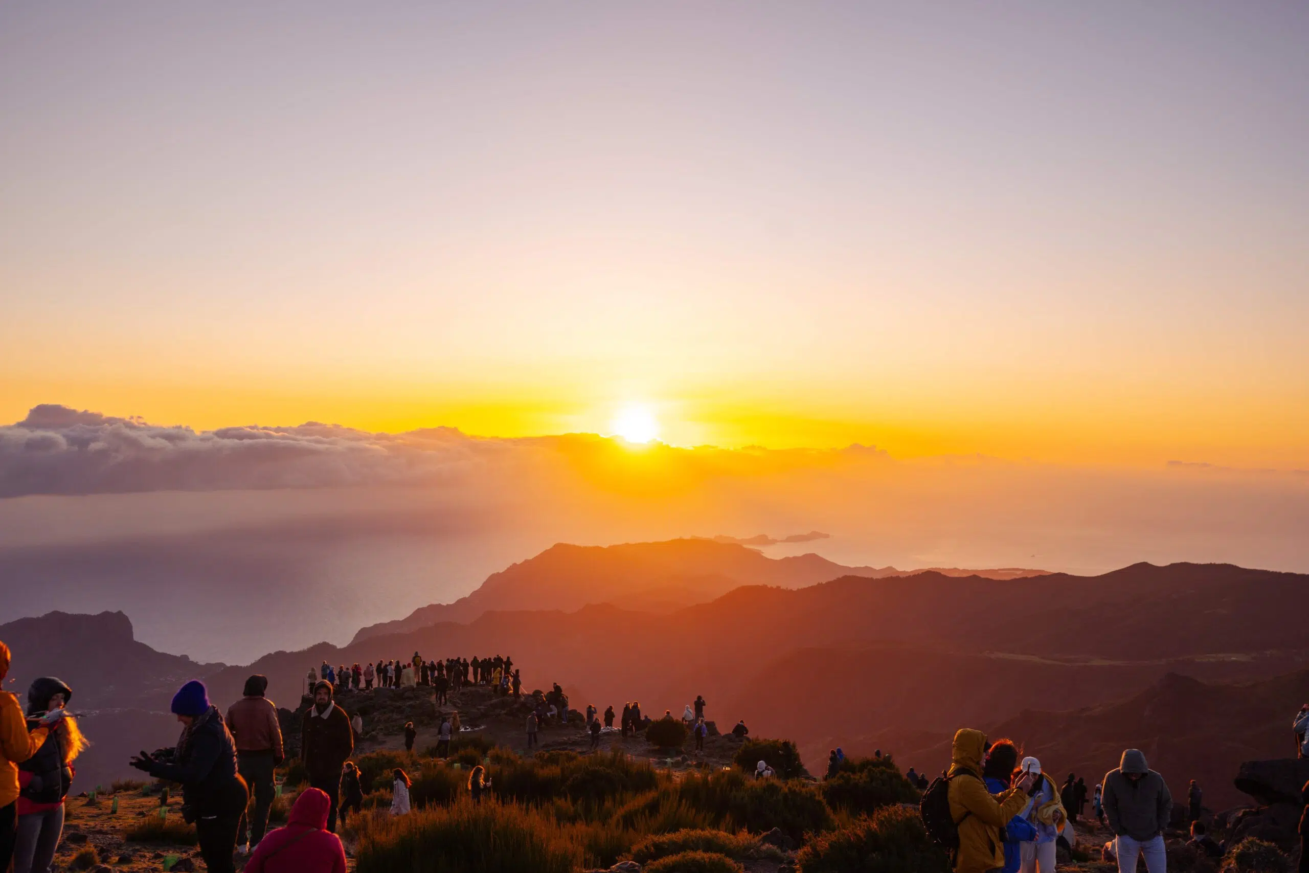 A group of people gather on a mountain peak at sunrise, silhouetted against a vivid orange and yellow sky. The mood is serene and awe-inspiring.