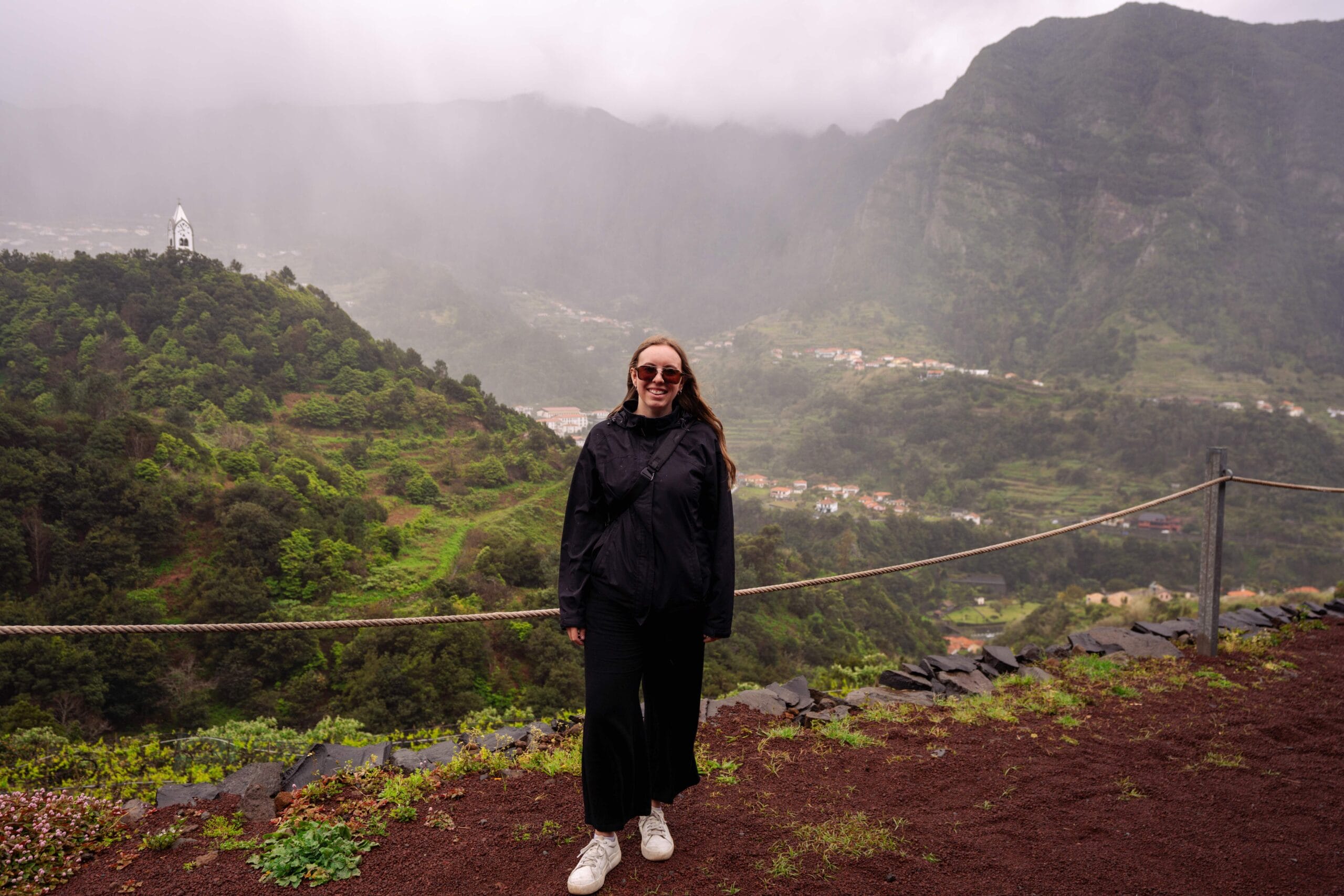 Woman in black jacket and sunglasses smiles, standing on a red dirt path with lush green hills and a foggy, mountainous landscape behind her. Peaceful mood.