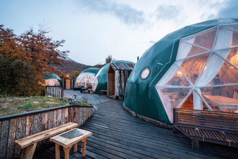 Geodesic domes with warm interior lighting sit on a wooden deck, surrounded by autumn trees under a cloudy sky, creating a cozy, eco-friendly atmosphere.