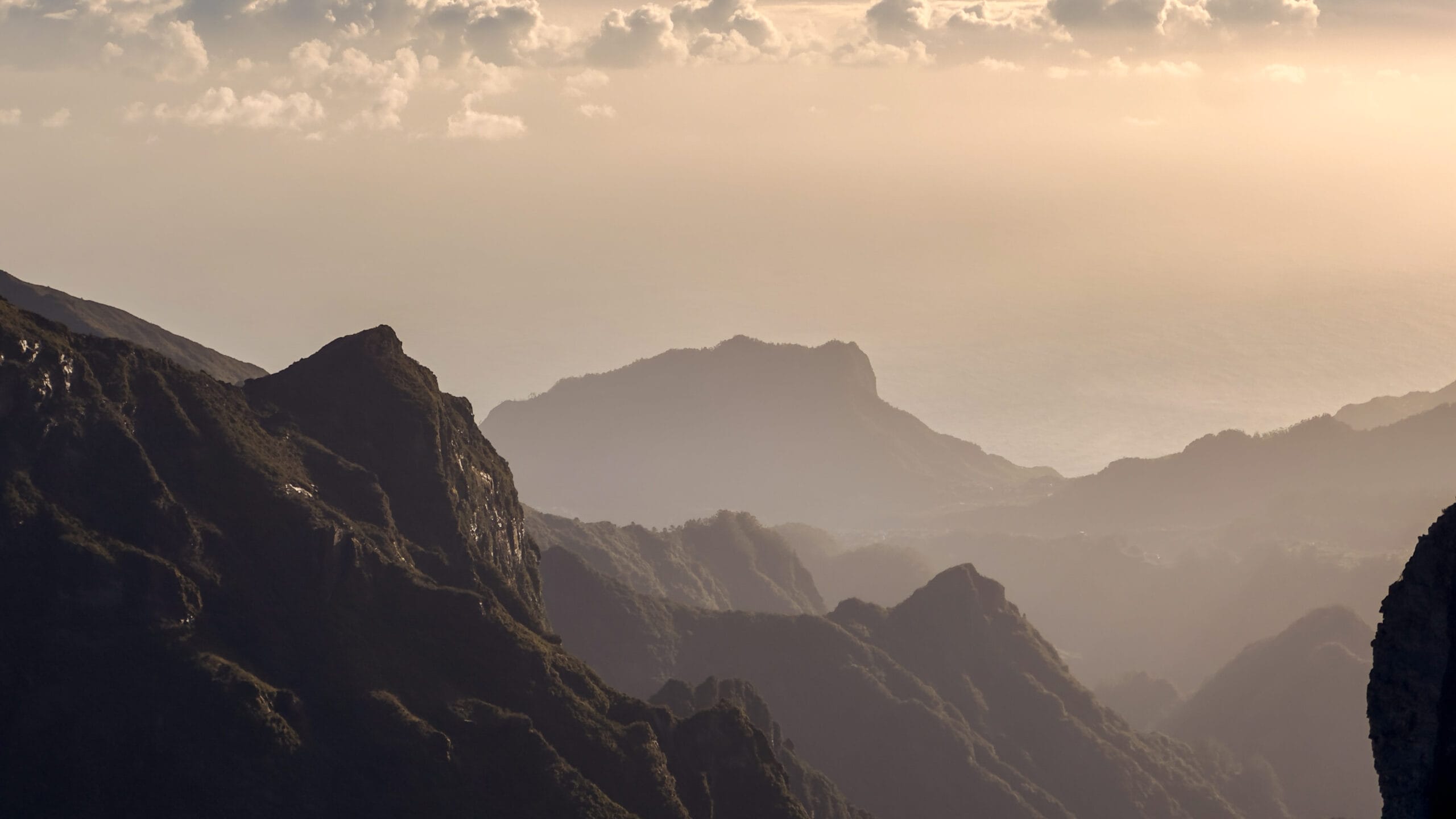 Misty mountain range at sunset with dark ridges in the foreground and softer, hazy peaks in the distance. A serene, tranquil atmosphere prevails.