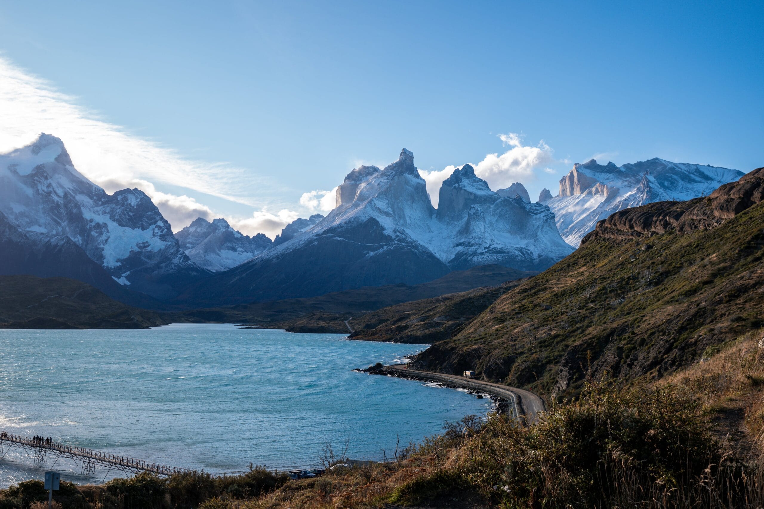Snow-capped mountains tower over a serene lake with a winding road and bridge in the foreground. The sky is clear and the scene is tranquil.