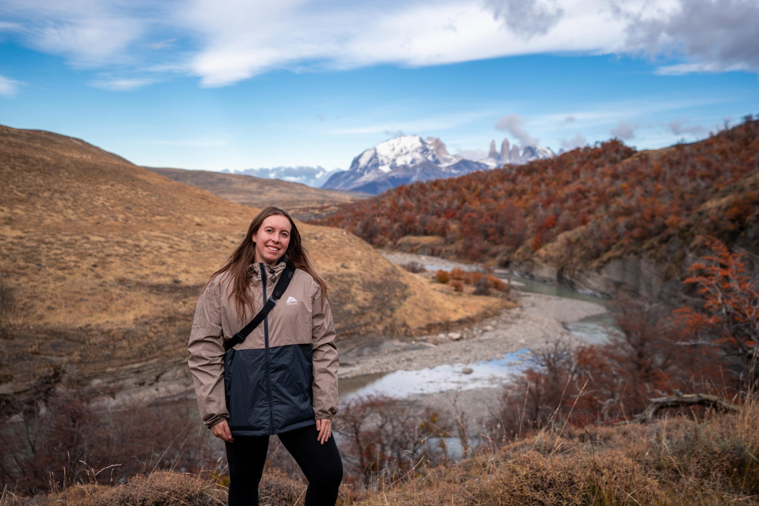 A woman in a beige and blue jacket smiles in a scenic landscape. Behind her, rolling hills, autumn trees, and snow-capped mountains under a blue sky.