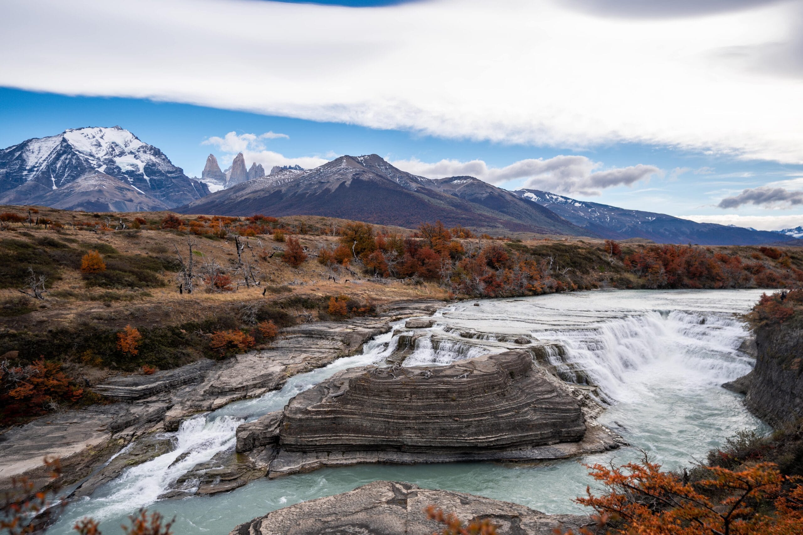 A scenic waterfall cascades over rocky ledges with snowy mountains in the background. Autumn foliage in orange and red adds warmth under a blue sky.