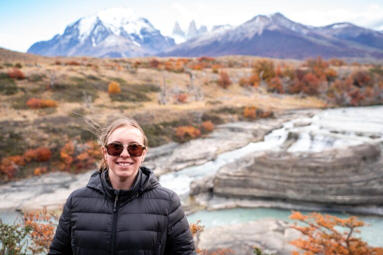 A smiling person in sunglasses and a black jacket stands in front of a scenic landscape with snowy mountains, autumn trees, and layered rock formations.
