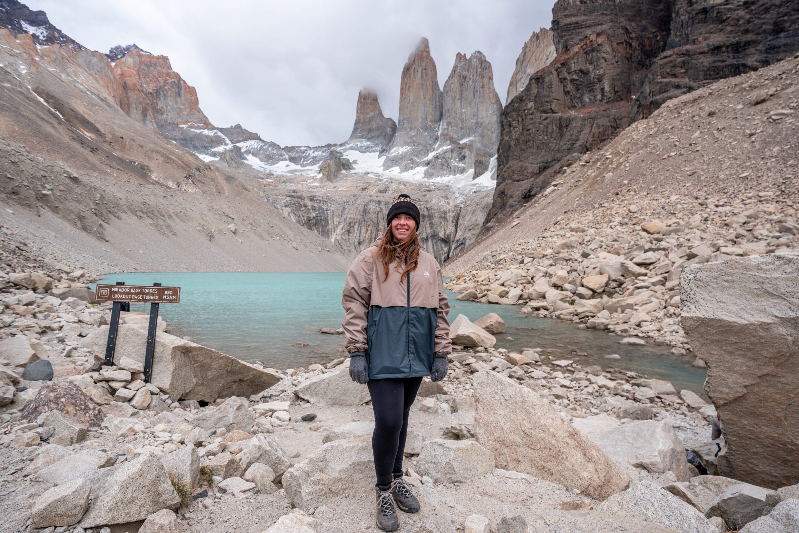 Smiling person in a winter jacket and beanie stands on rocky terrain near turquoise lake, with towering snow-capped peaks and cloudy sky in the background.
