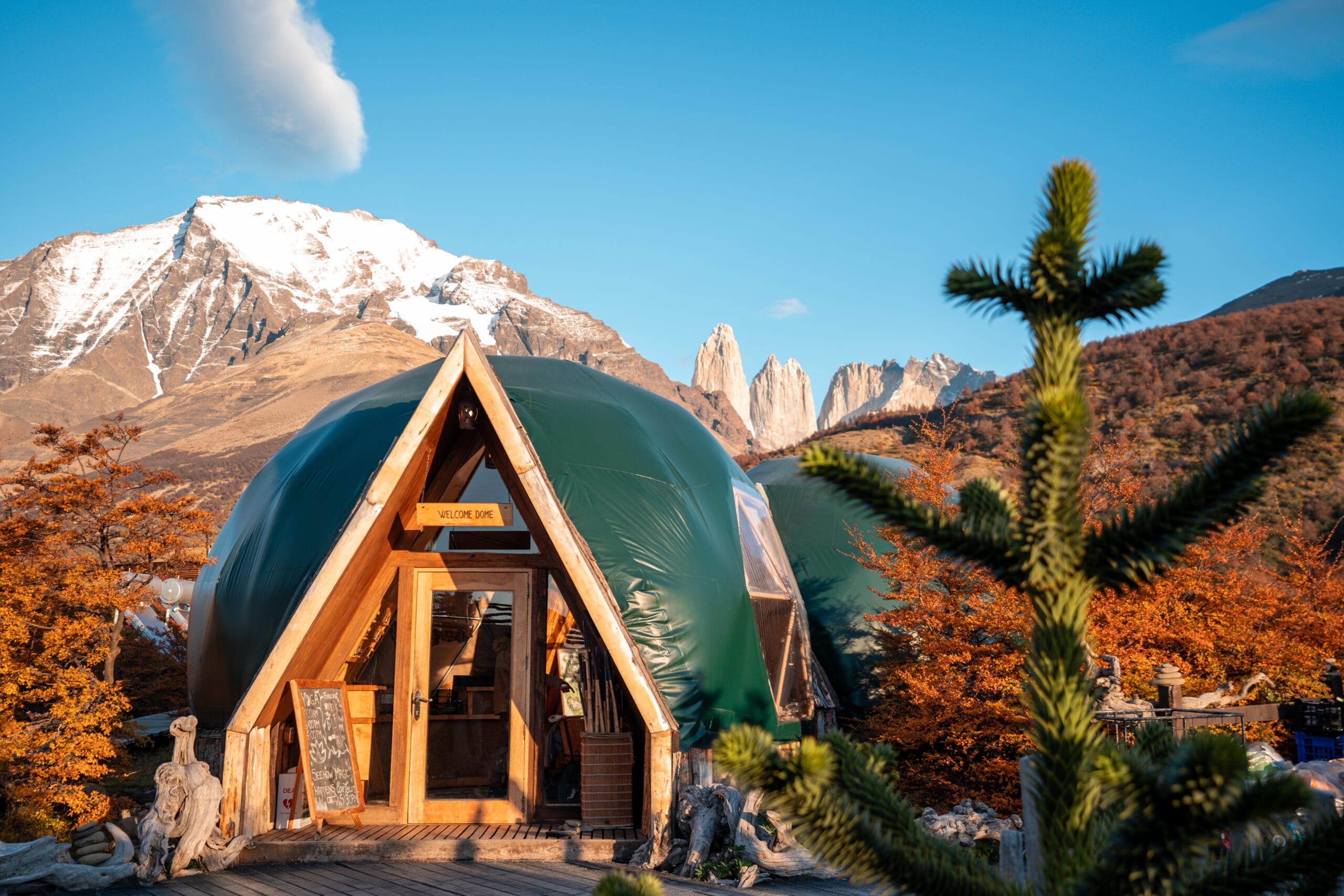 Dome-shaped cabin with wooden entrance set in autumn-colored forest, framed by snow-capped mountains under a clear blue sky, conveying tranquility.