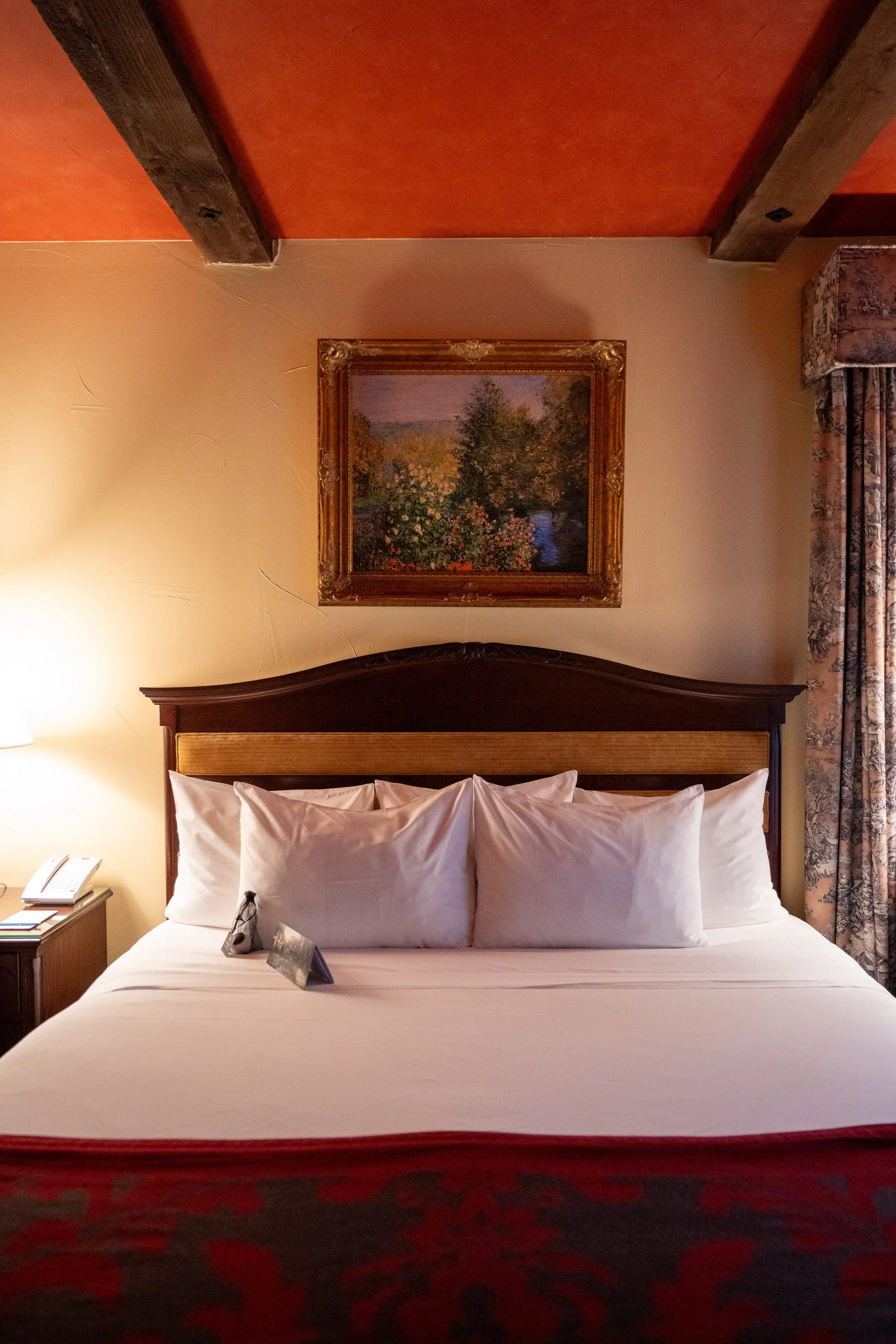 Cozy hotel room with a wooden bed, four white pillows, a red and gray patterned throw. Warm lighting, landscape painting above headboard.