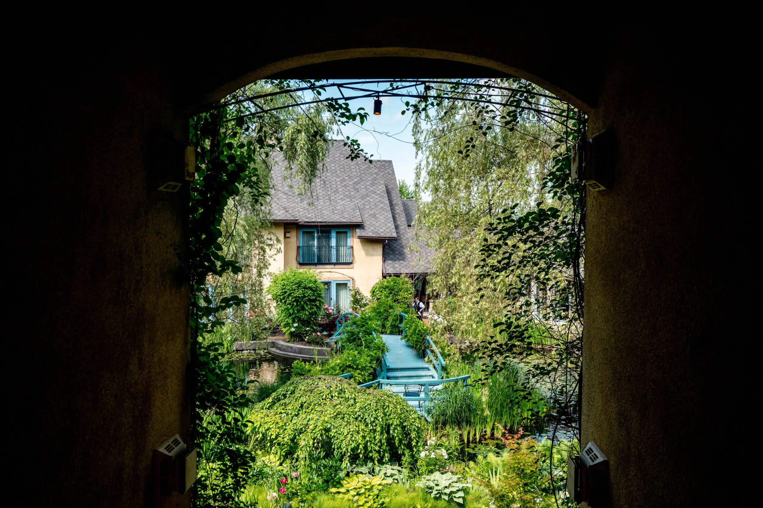 A cozy house with a sloped roof is framed by an arched doorway, surrounded by lush greenery and trees. A tranquil blue bridge crosses a serene garden.