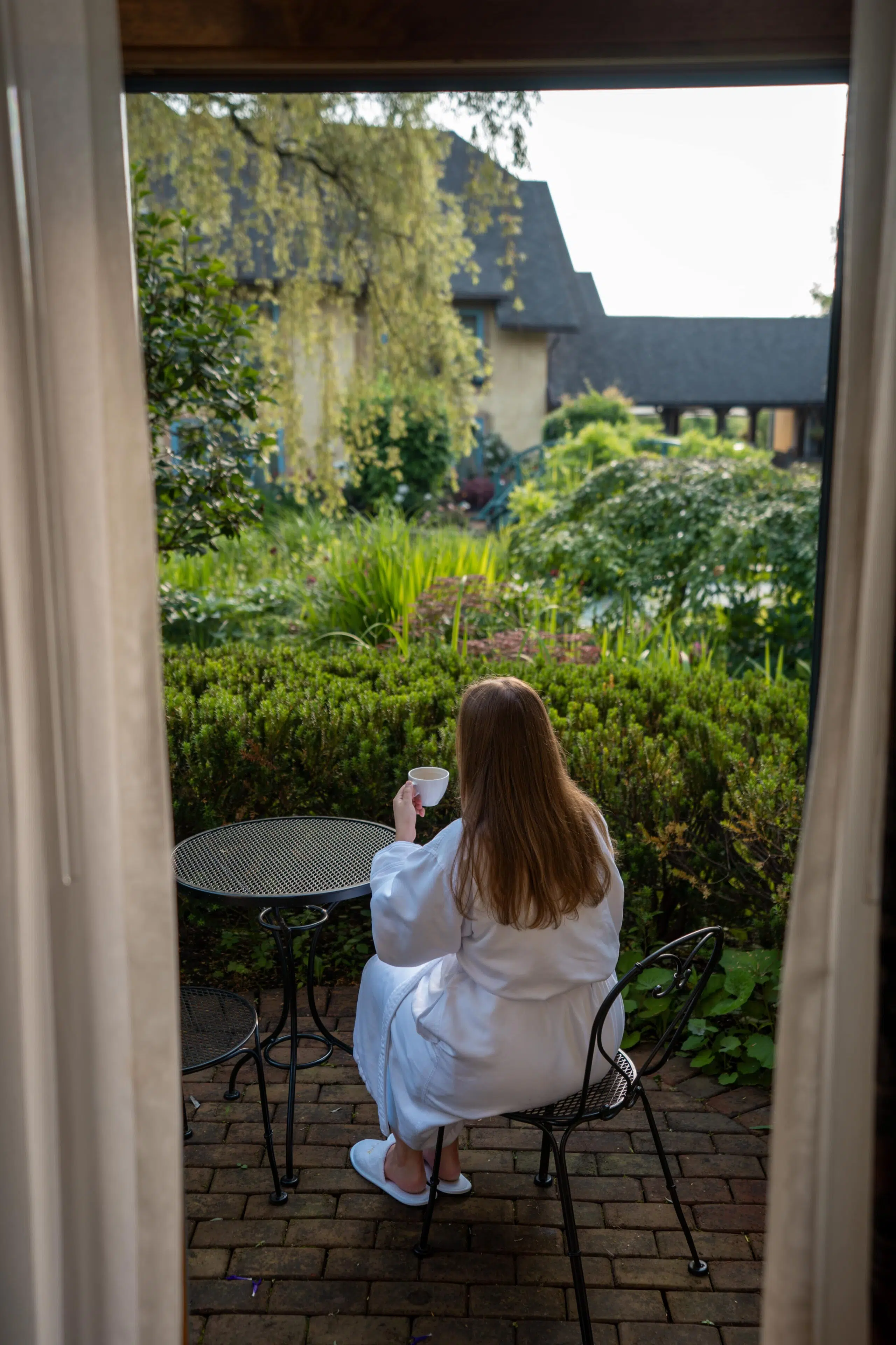 A woman in a white robe sits on a patio chair, holding a cup, overlooking a lush garden and cottage. The scene conveys tranquility and relaxation.