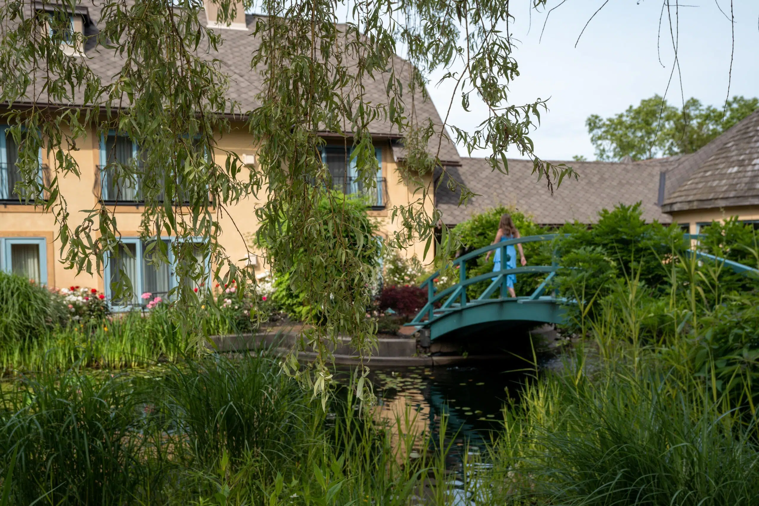 Charming garden scene with a picturesque bridge over a pond, lush greenery, and a quaint house in the background. A person strolls at mirbeau spa skaneateles on the bridge.