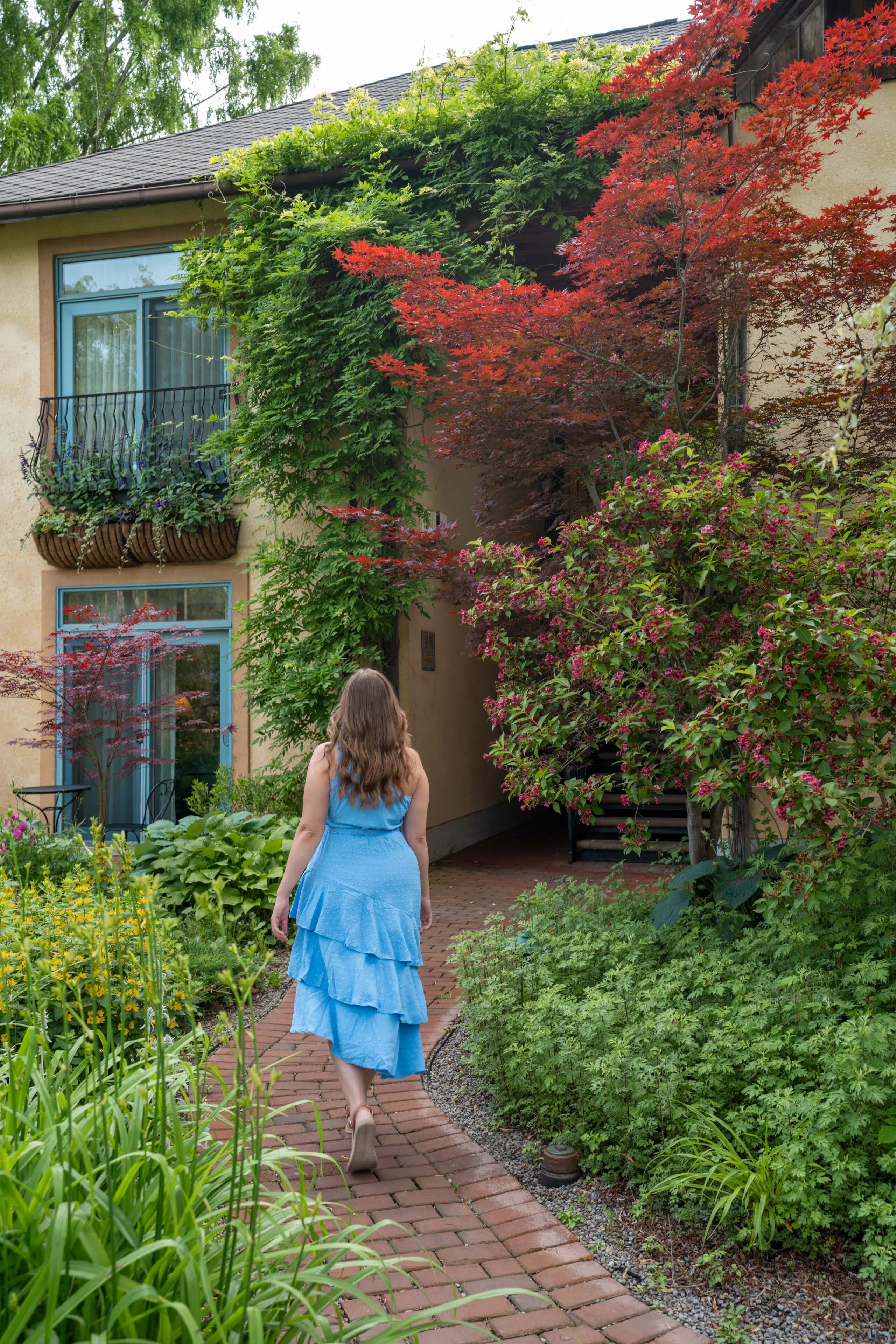 A woman in a blue dress walks along a brick path in a lush garden, surrounded by green and red foliage, approaching a charming, plant-covered house.