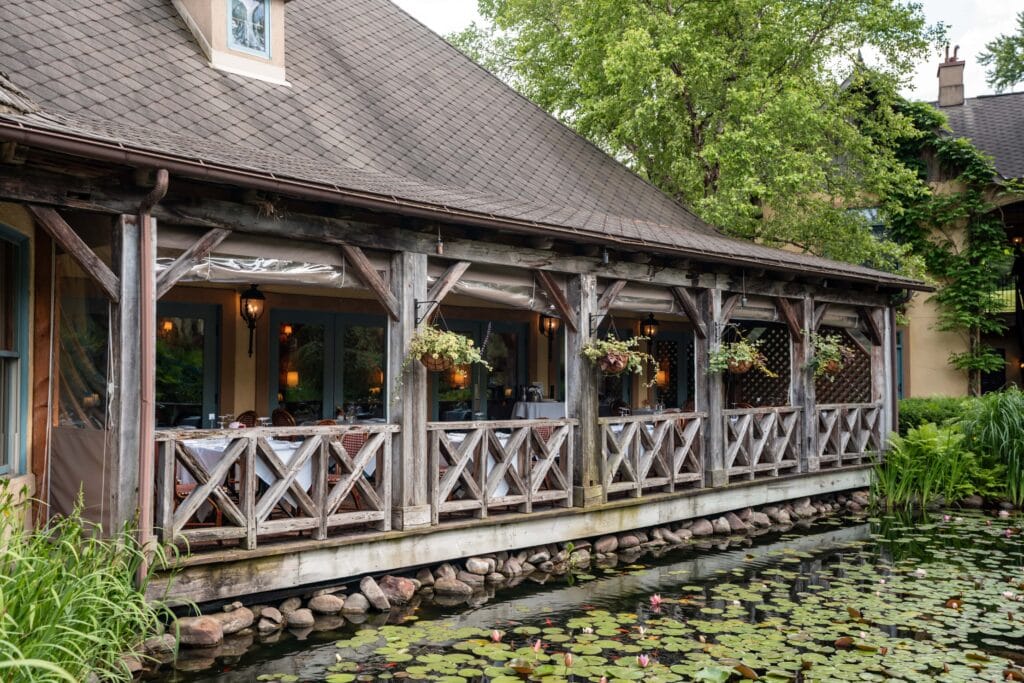 Rustic outdoor restaurant patio with wooden railing overlooks a pond filled with lily pads. Hanging plants and serene, lush greenery create a tranquil vibe at Mirbeau Spa Skaneateles.