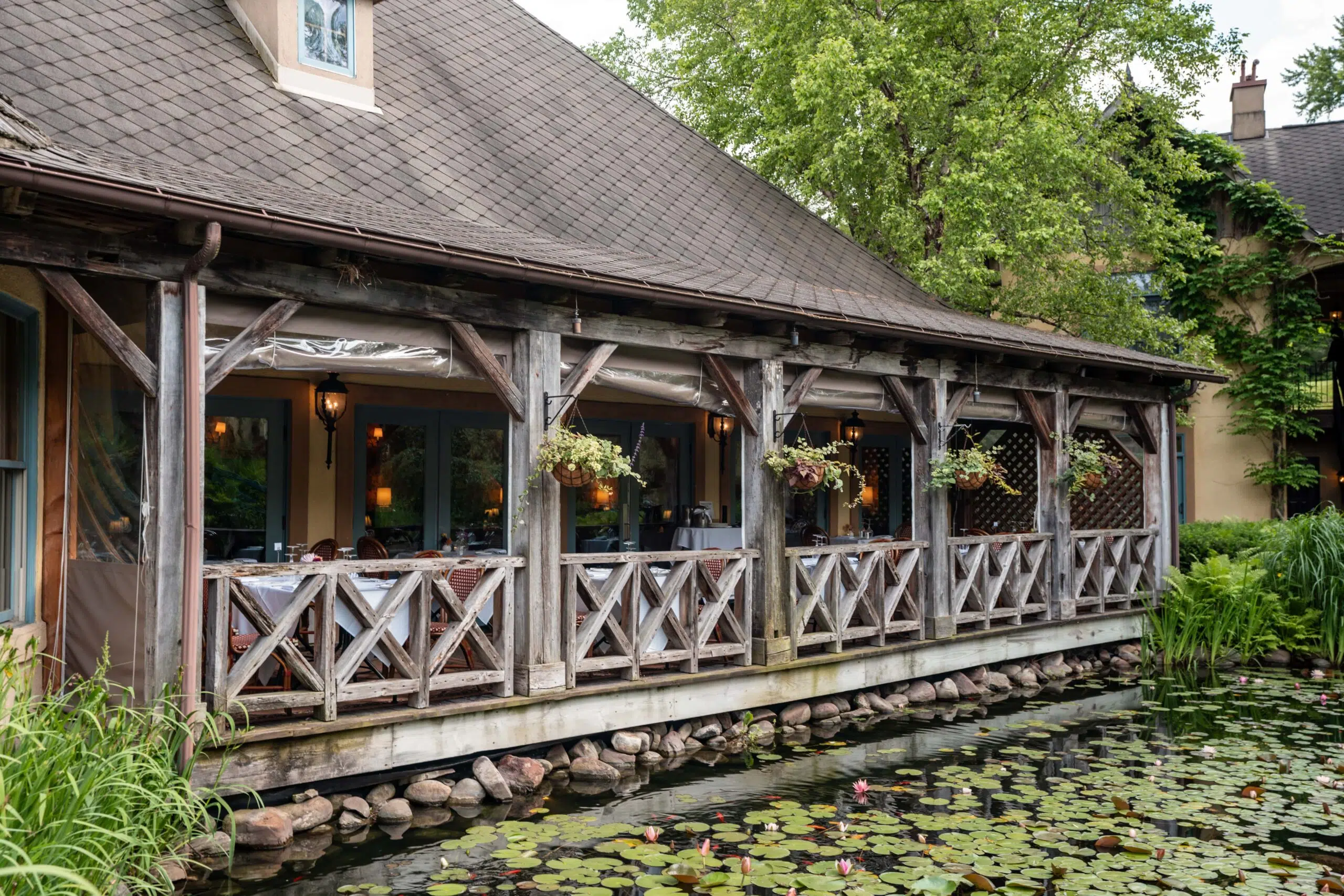 A rustic, wooden restaurant patio with hanging plants overlooks a serene pond filled with lily pads. The setting conveys a peaceful, rustic ambiance.