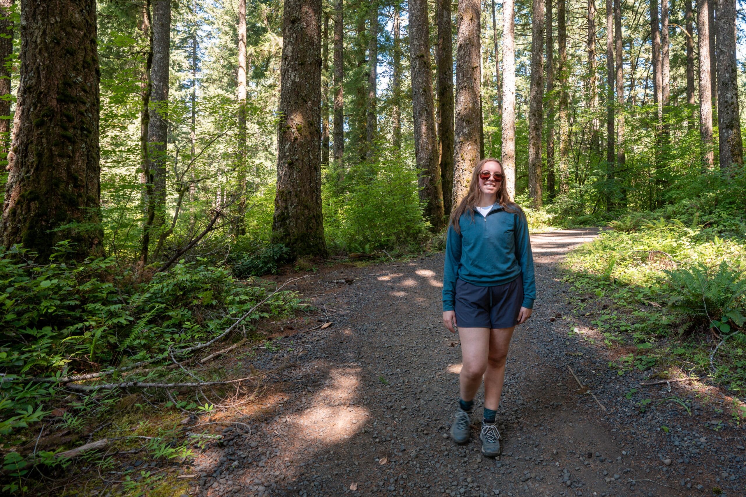 A woman wearing sunglasses, a blue long-sleeve shirt, and shorts stands smiling on a sunlit forest trail surrounded by tall trees and greenery.