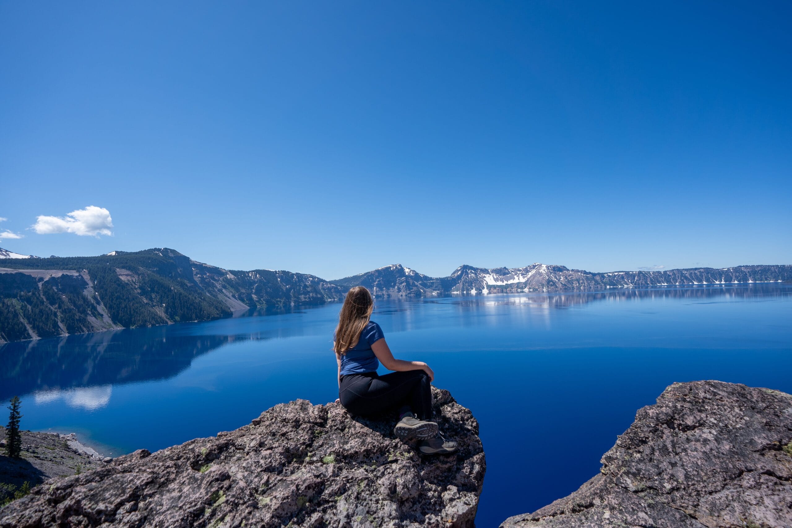 A woman sits on a rocky cliff, gazing at a vast, calm blue lake surrounded by mountains under a clear blue sky, conveying peace and contemplation.