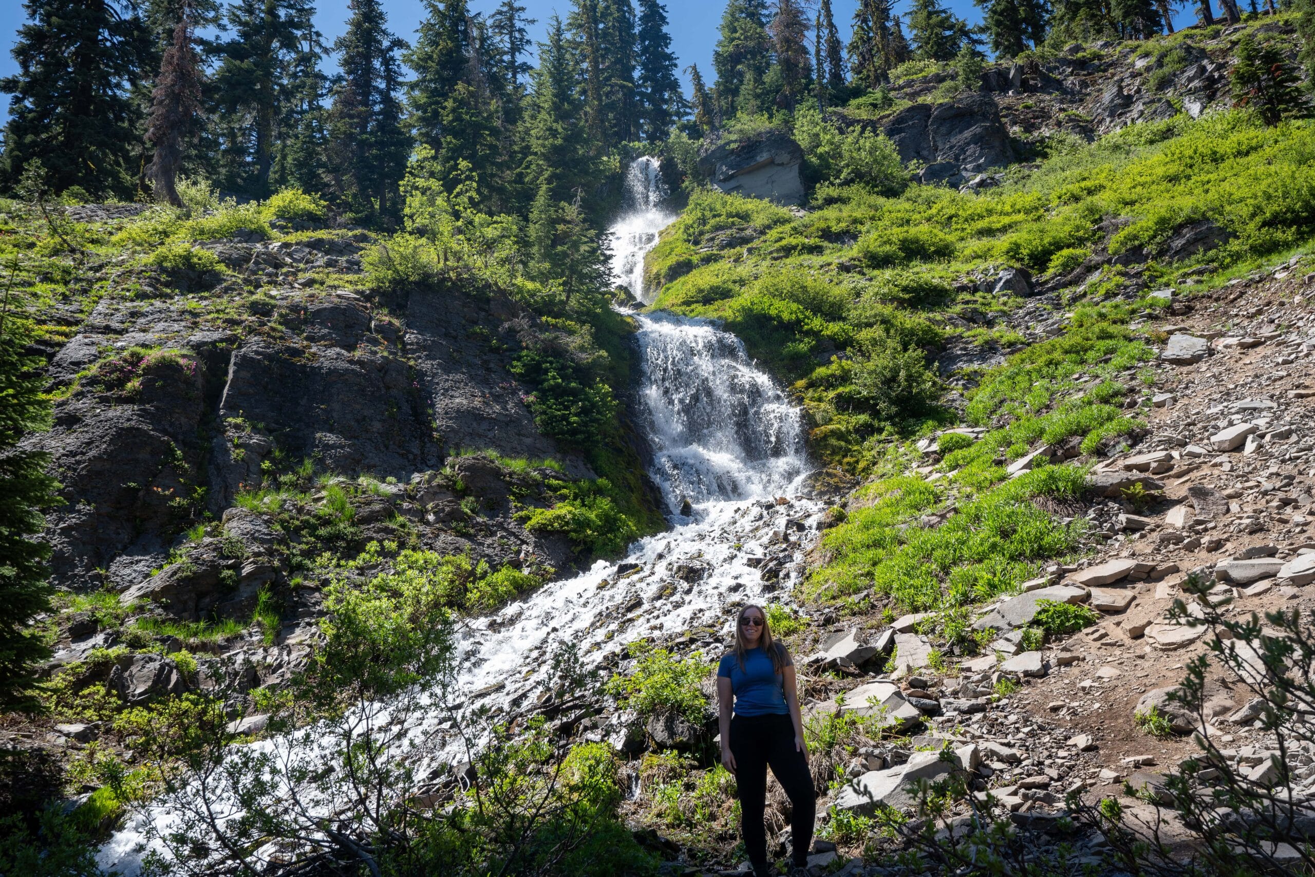 A woman stands near a cascading waterfall on a rocky hillside. The scene is lush and green, surrounded by tall trees, under a clear blue sky.