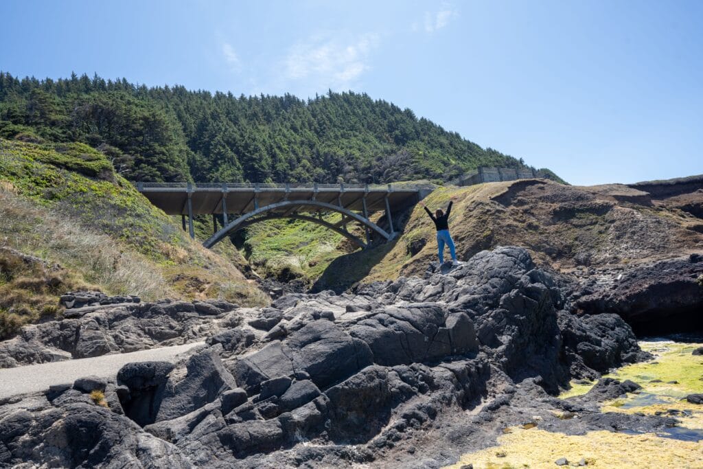 Person raising arms in merino wool in triumph atop rocky terrain near a large arched bridge. Dense green pine forest in the background under a clear blue sky.