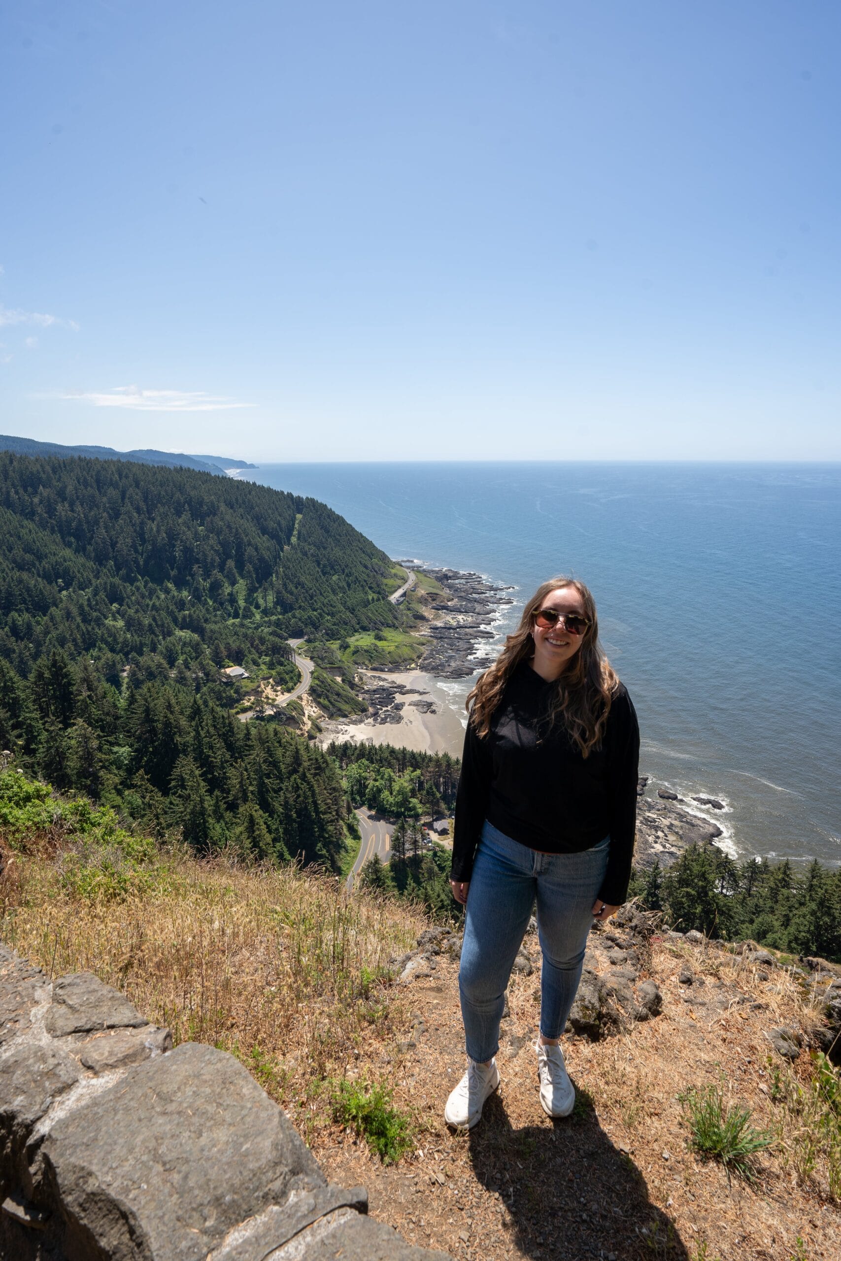 A person in sunglasses stands on a cliff overlooking a scenic coastline with lush green forests and the ocean under a clear blue sky.