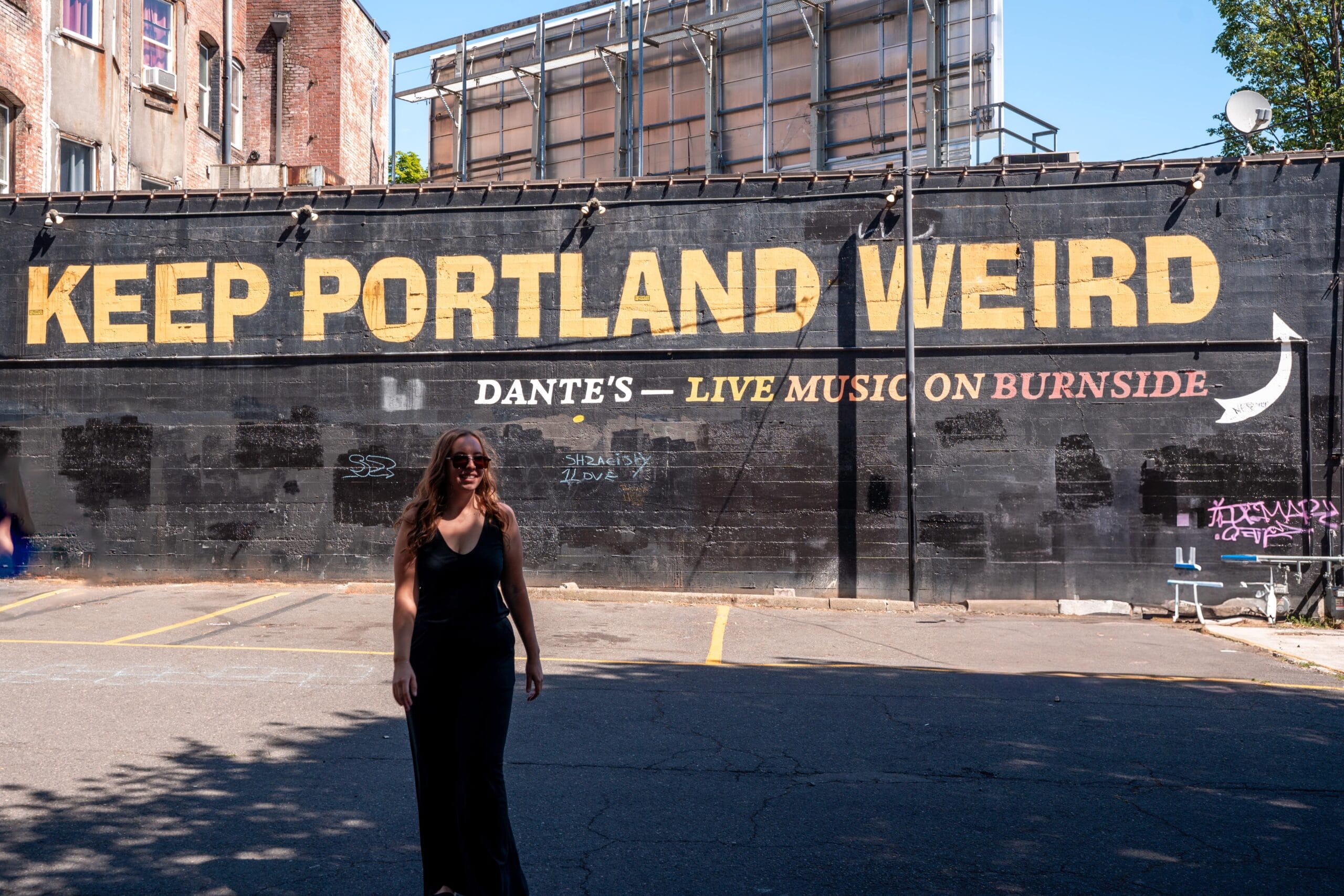 A woman in a black dress stands smiling wearing black merino wool in front of a brick wall with the slogan "KEEP PORTLAND WEIRD" in bold yellow text, conveying a quirky, cheerful vibe.
