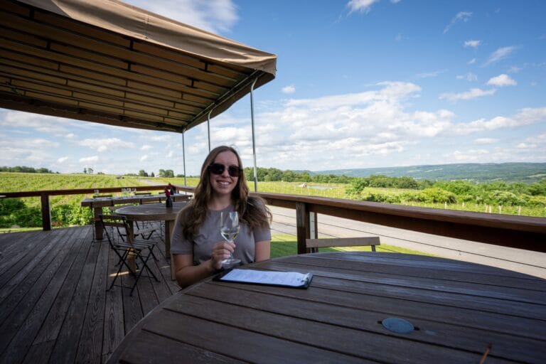 A woman wearing the best merino wool travel clothes and sunglasses sits at a wooden table on a sunny patio, holding a glass of wine. Lush vineyards and a clear blue sky are in the background.