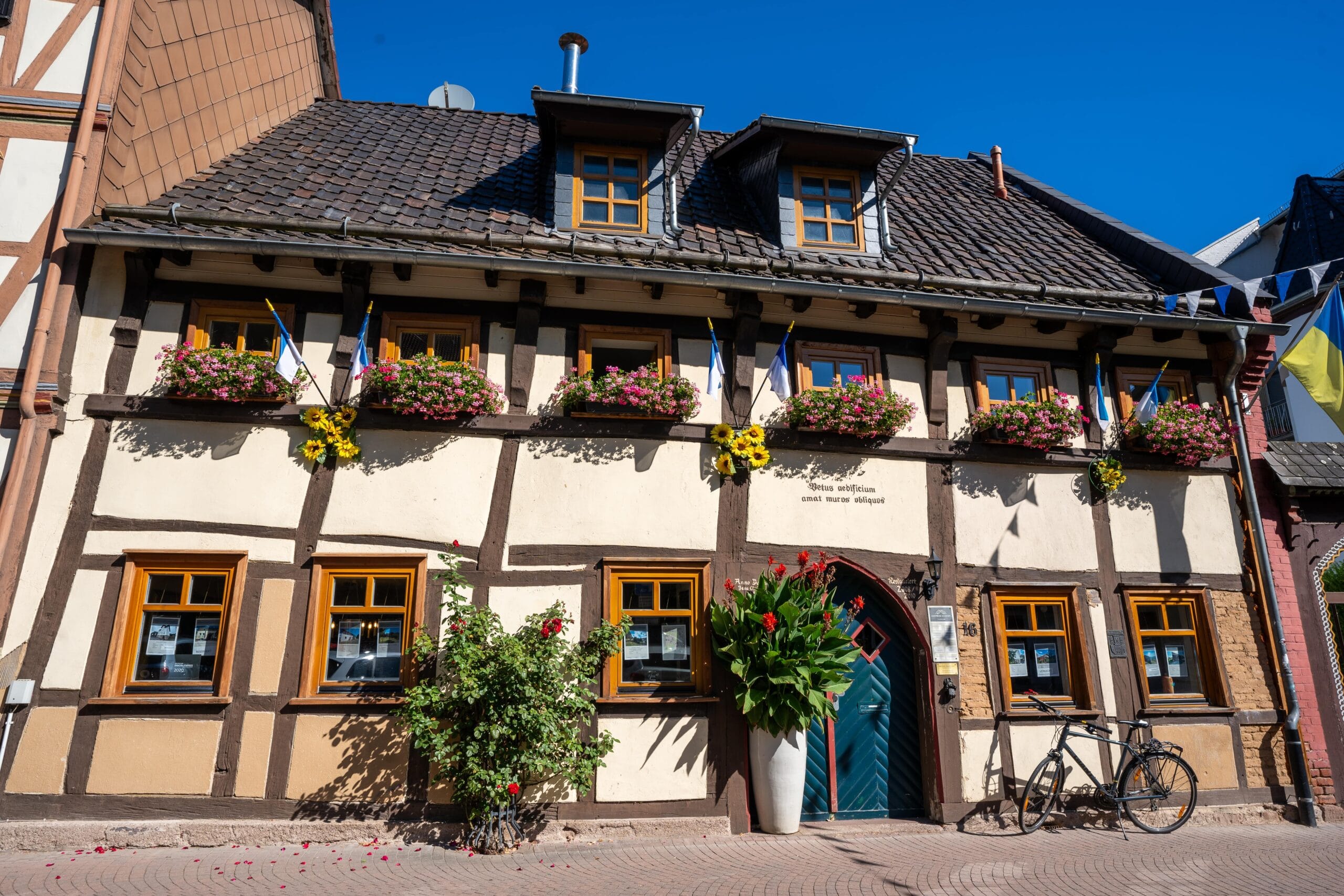Half-timbered house with colorful flowers in window boxes and flags, under a clear blue sky. A bicycle and potted plants add charm and vibrancy.