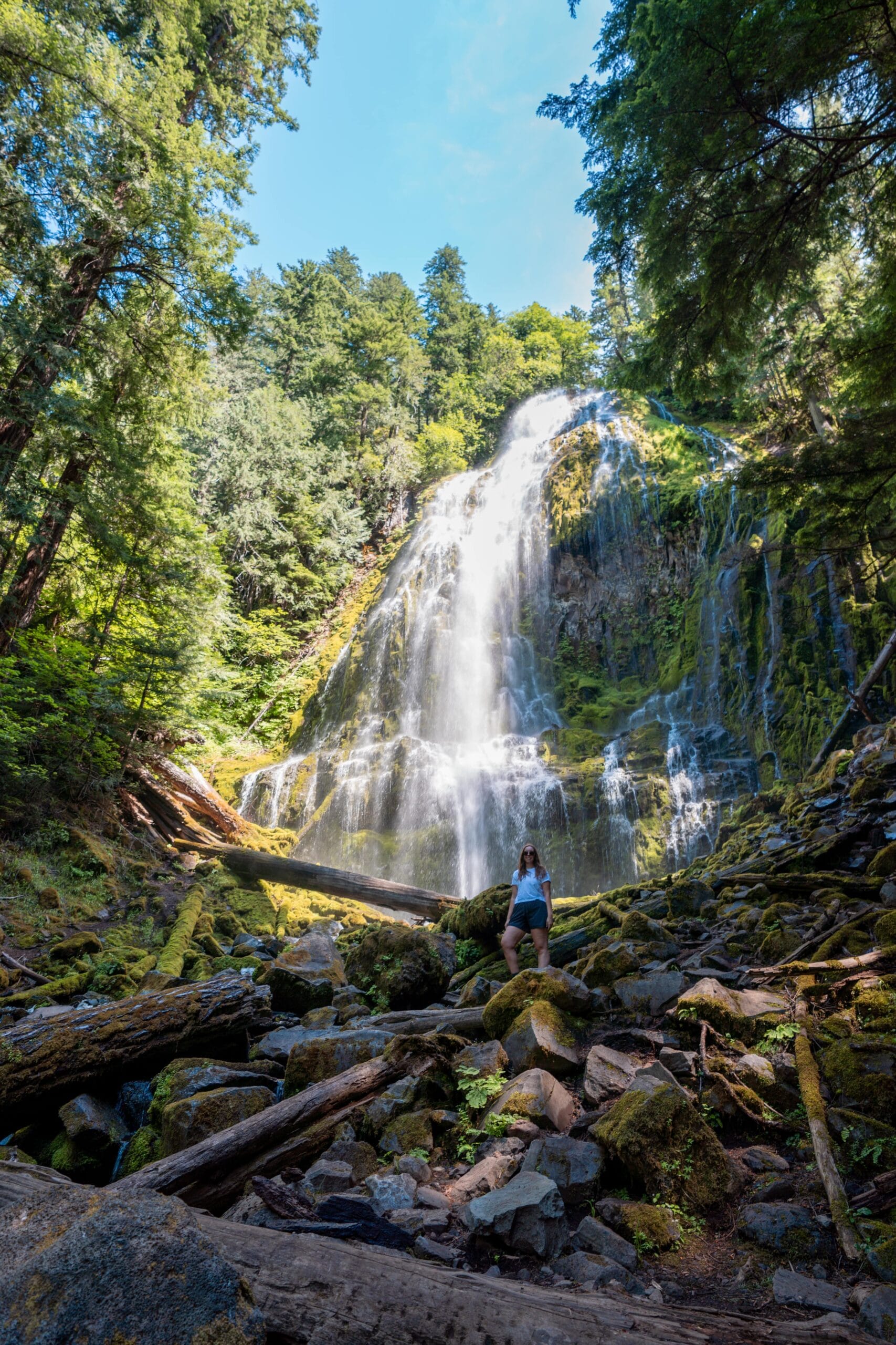 A woman stands on mossy rocks at the base of a tall, cascading waterfall surrounded by lush green forest. Sunlight filters through the trees, creating a serene and majestic atmosphere.