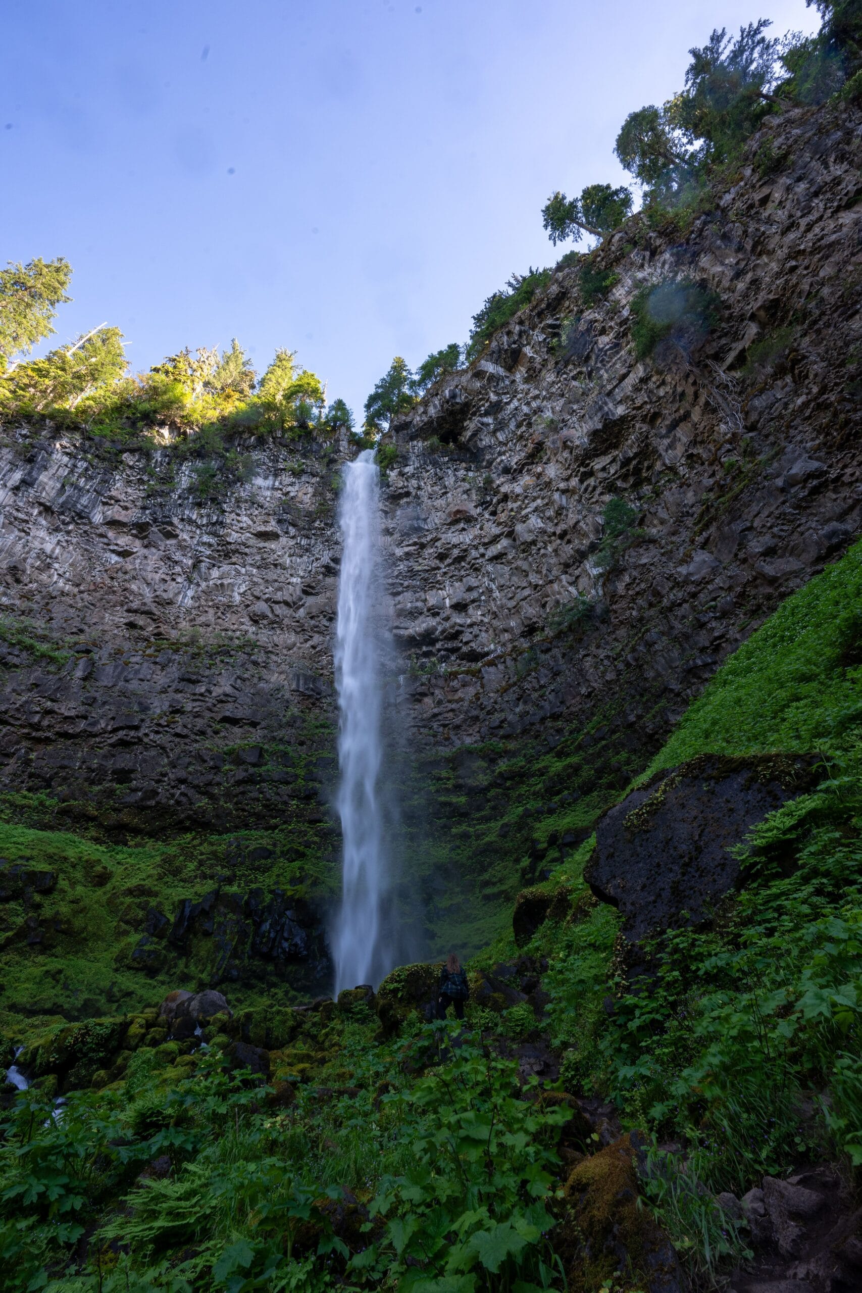Tall waterfall cascading down a rocky cliff surrounded by lush greenery. A small figure stands at the base, arms raised, conveying awe and wonder.