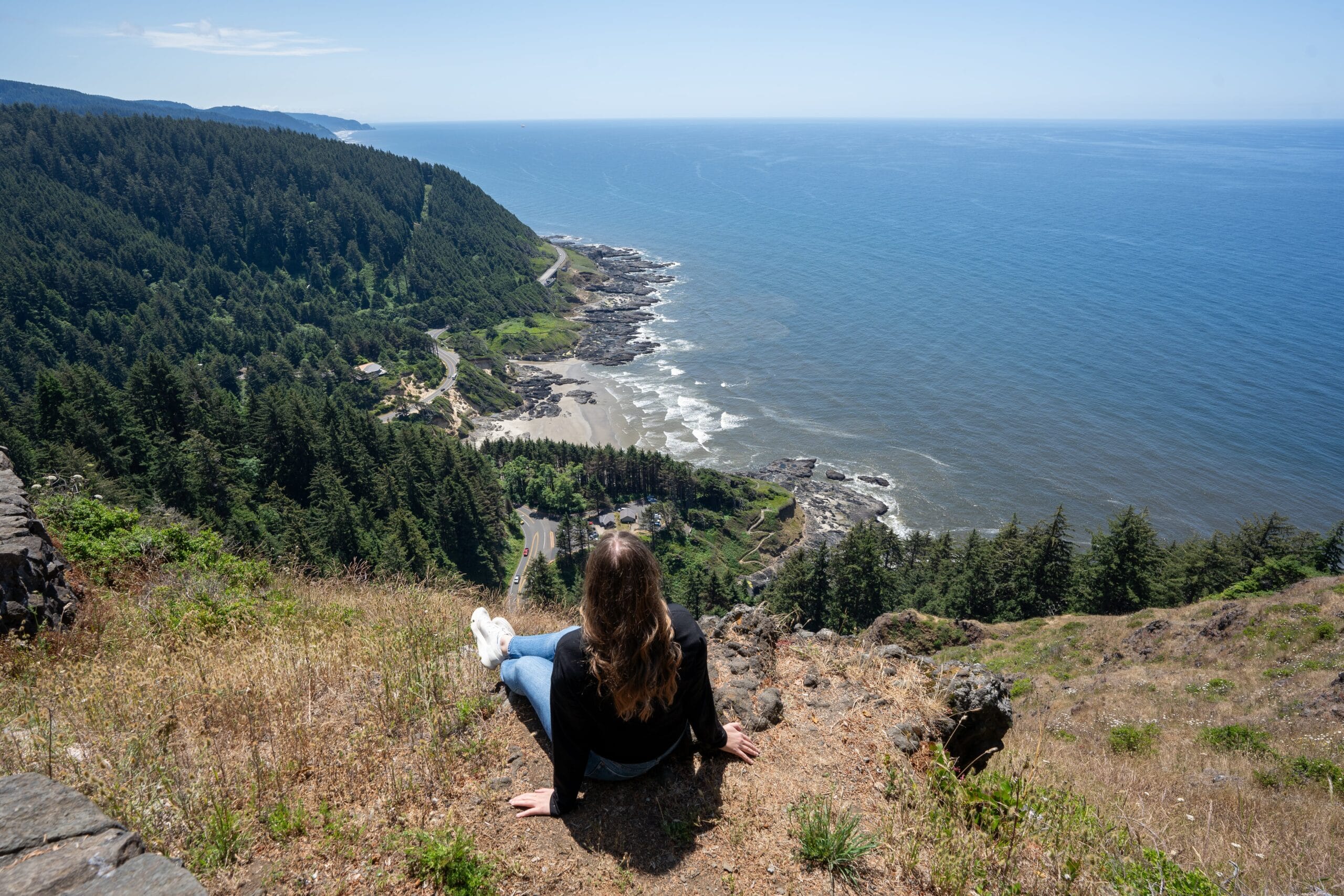 A woman with long hair sits on a grassy cliff, overlooking a vast ocean and rocky shoreline. The scene is peaceful with a sunny, clear blue sky.