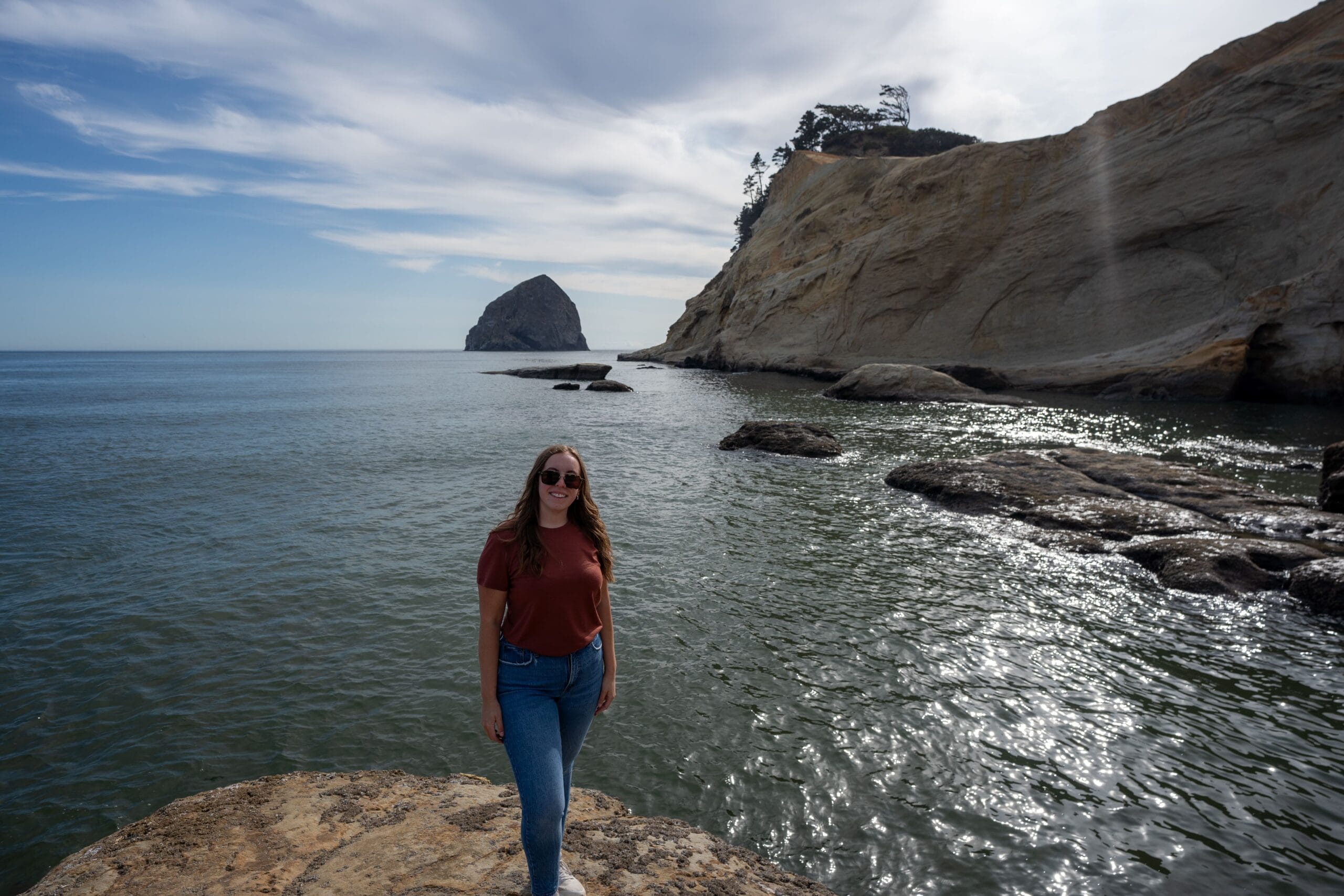 A woman in sunglasses and casual attire stands smiling on a rocky shore. The sea sparkles under a partly cloudy sky; a large rock formation is in the distance.