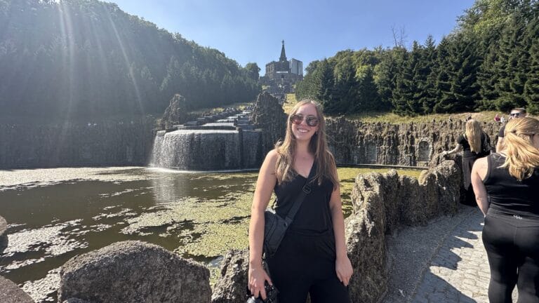 A smiling woman in sunglasses stands beside a rocky pathway overlooking a waterfall and pond, with a towering forest and majestic castle in the background, under a clear blue sky.
