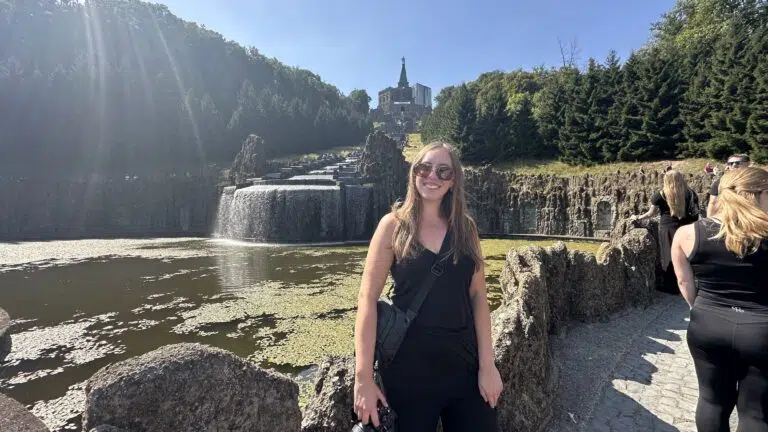 A smiling woman in sunglasses stands beside a rocky pathway overlooking a waterfall and pond, with a towering forest and majestic castle in the background, under a clear blue sky.
