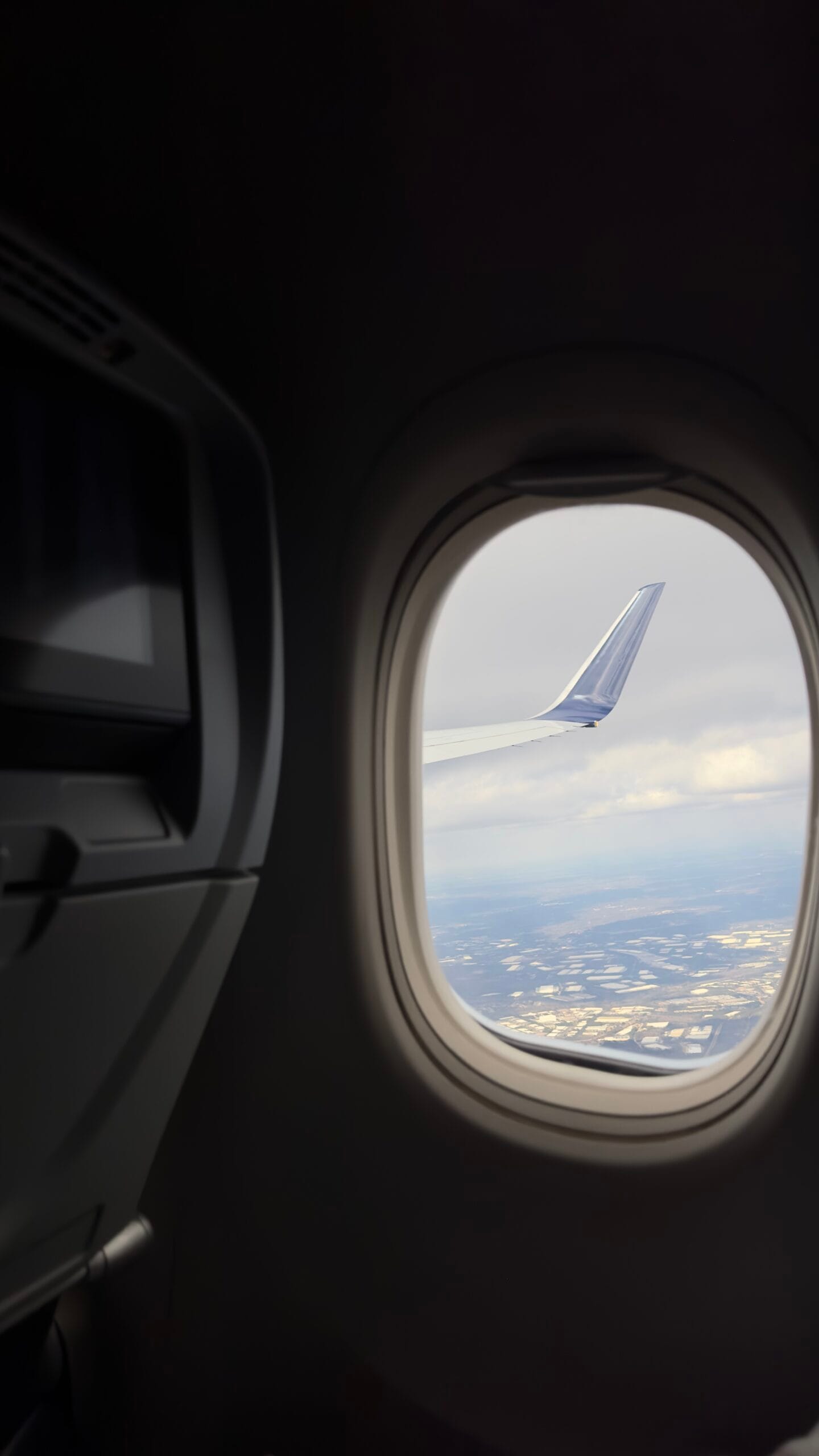 View from an airplane window showing a wing against a cloudy sky, with a glimpse of land below. The cabin interior is dimly lit, creating a serene atmosphere.