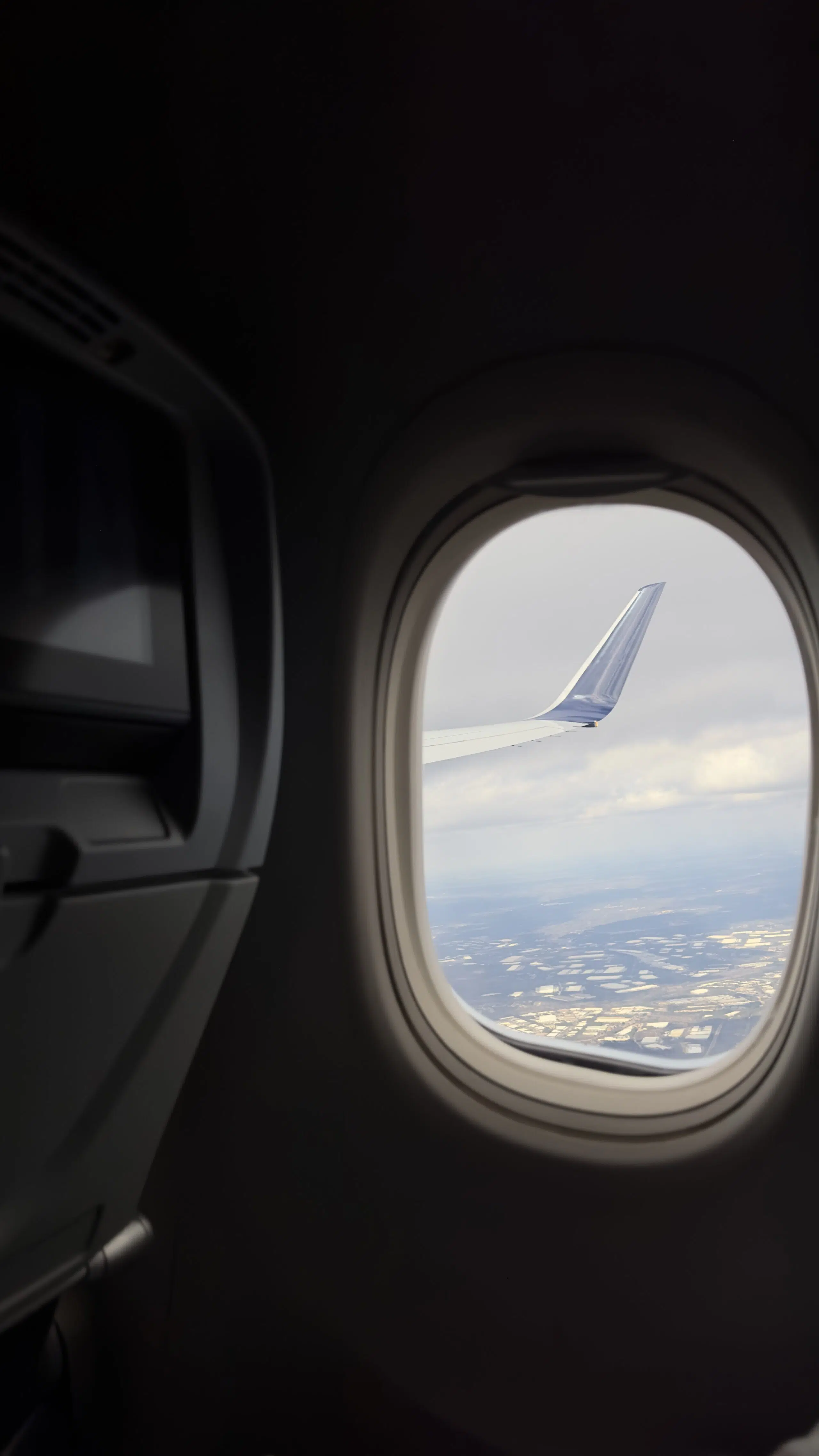 View from an airplane window showing a wing against a cloudy sky, with a glimpse of land below. The cabin interior is dimly lit, creating a serene atmosphere.