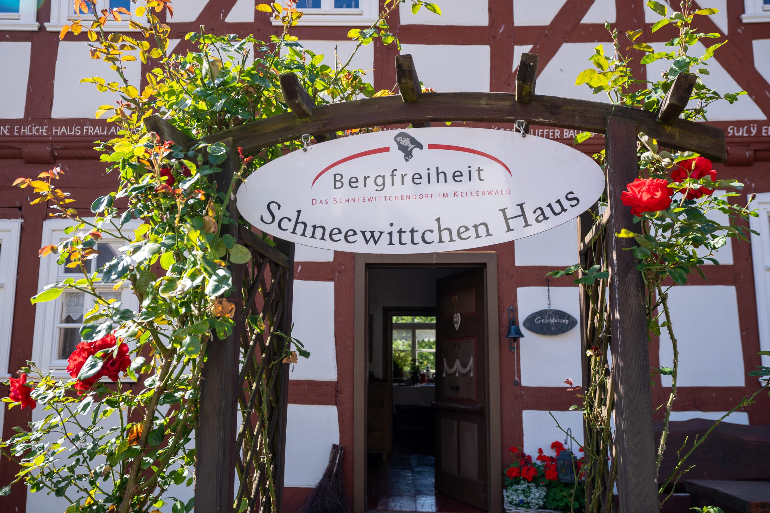 Wooden arbor with red roses and a sign reading "Schneewittchen Haus" leads to a quaint half-timbered house. Bright and inviting atmosphere.