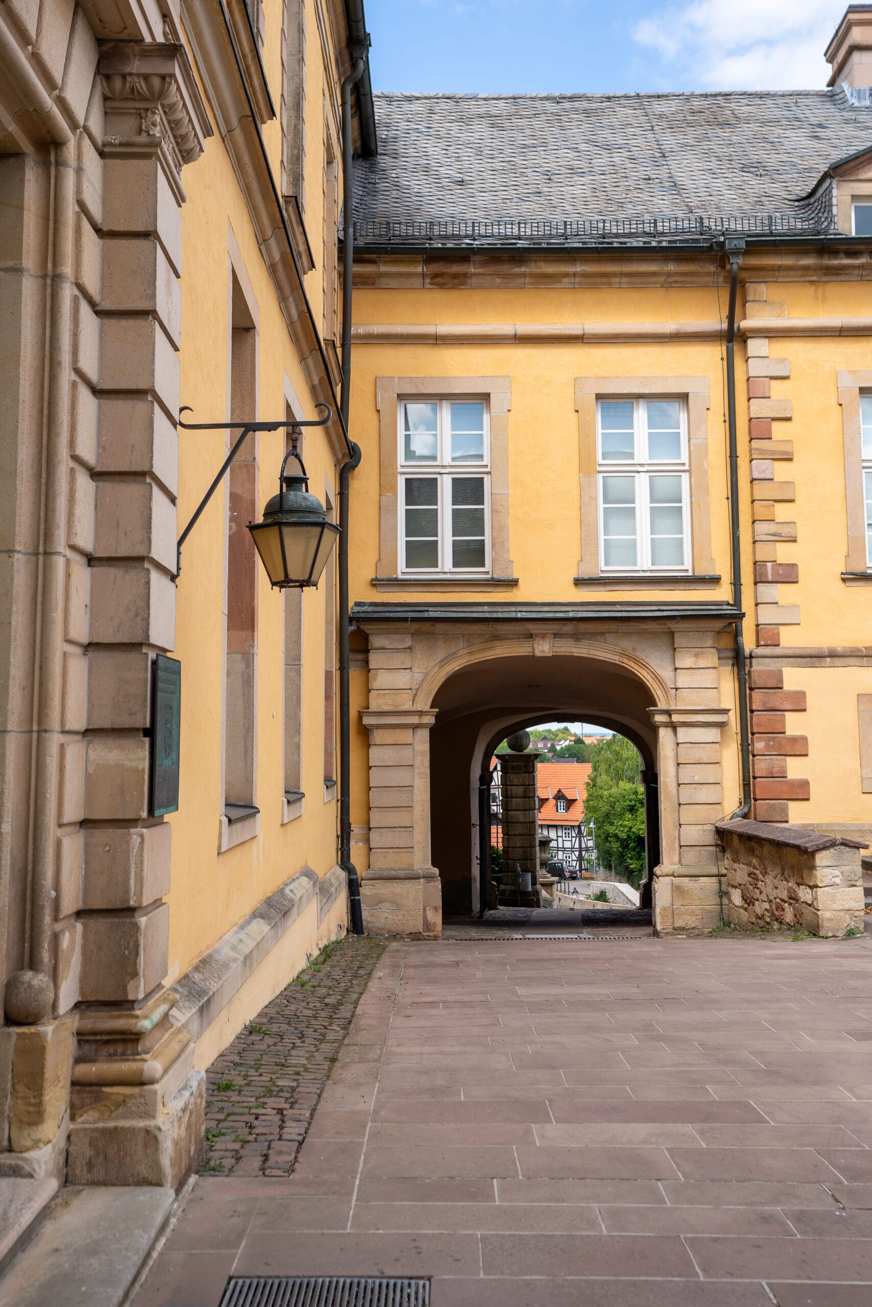 A charming yellow building with arched walkway leads to a scenic view of rooftops and greenery. Warm tones, historic architecture, peaceful ambiance.