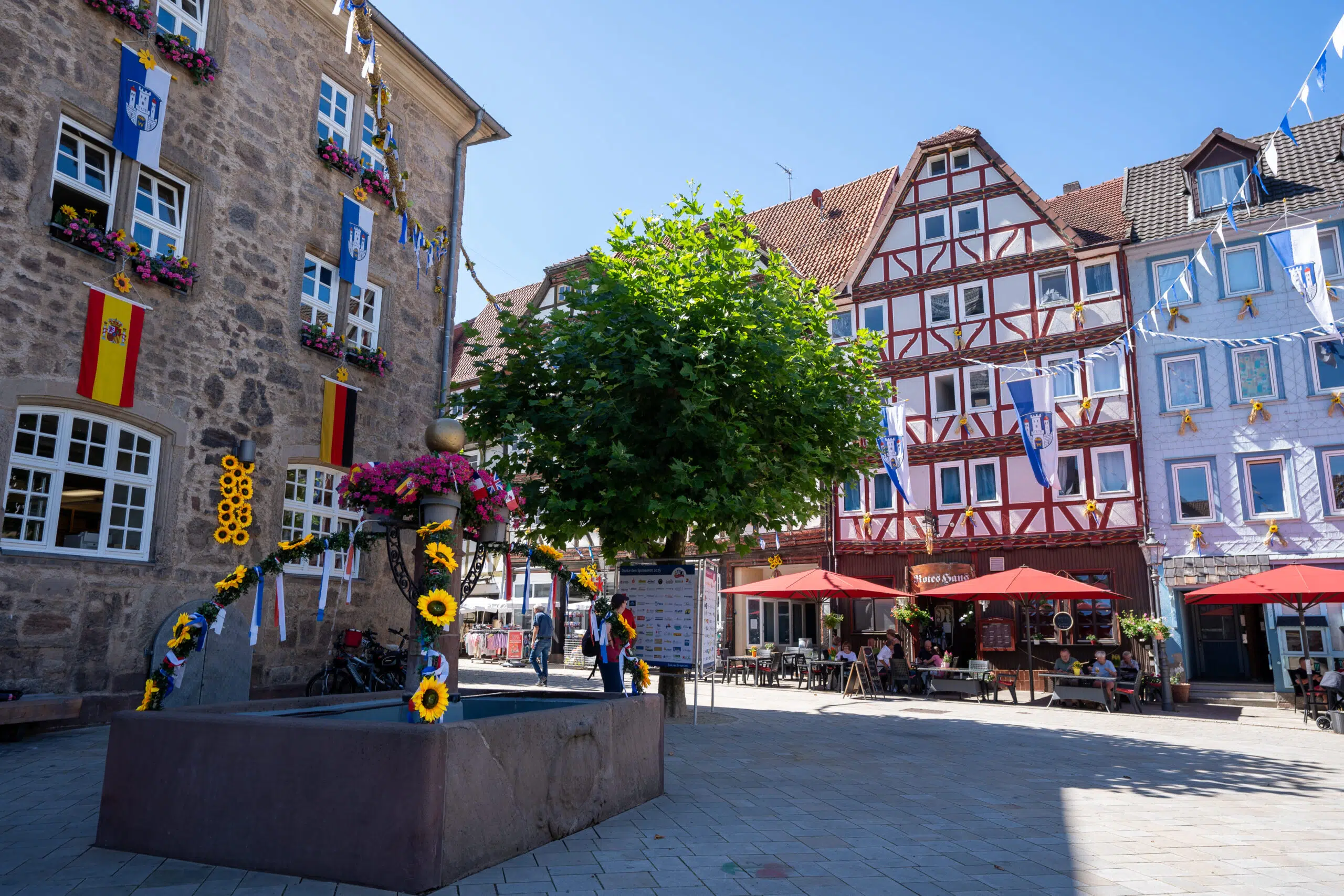 A charming fairy tale route in germany square under a clear blue sky, featuring a decorated fountain with sunflowers, historic buildings adorned with flags, and outdoor cafes.