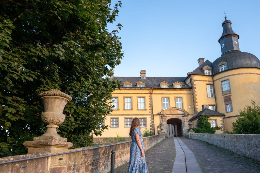 A woman in a blue dress stands on a cobblestone path leading to a historic yellow castle with rounded turret. A large tree and stone urn are on the left. In Bad Wildungen during a Germany itinerary.