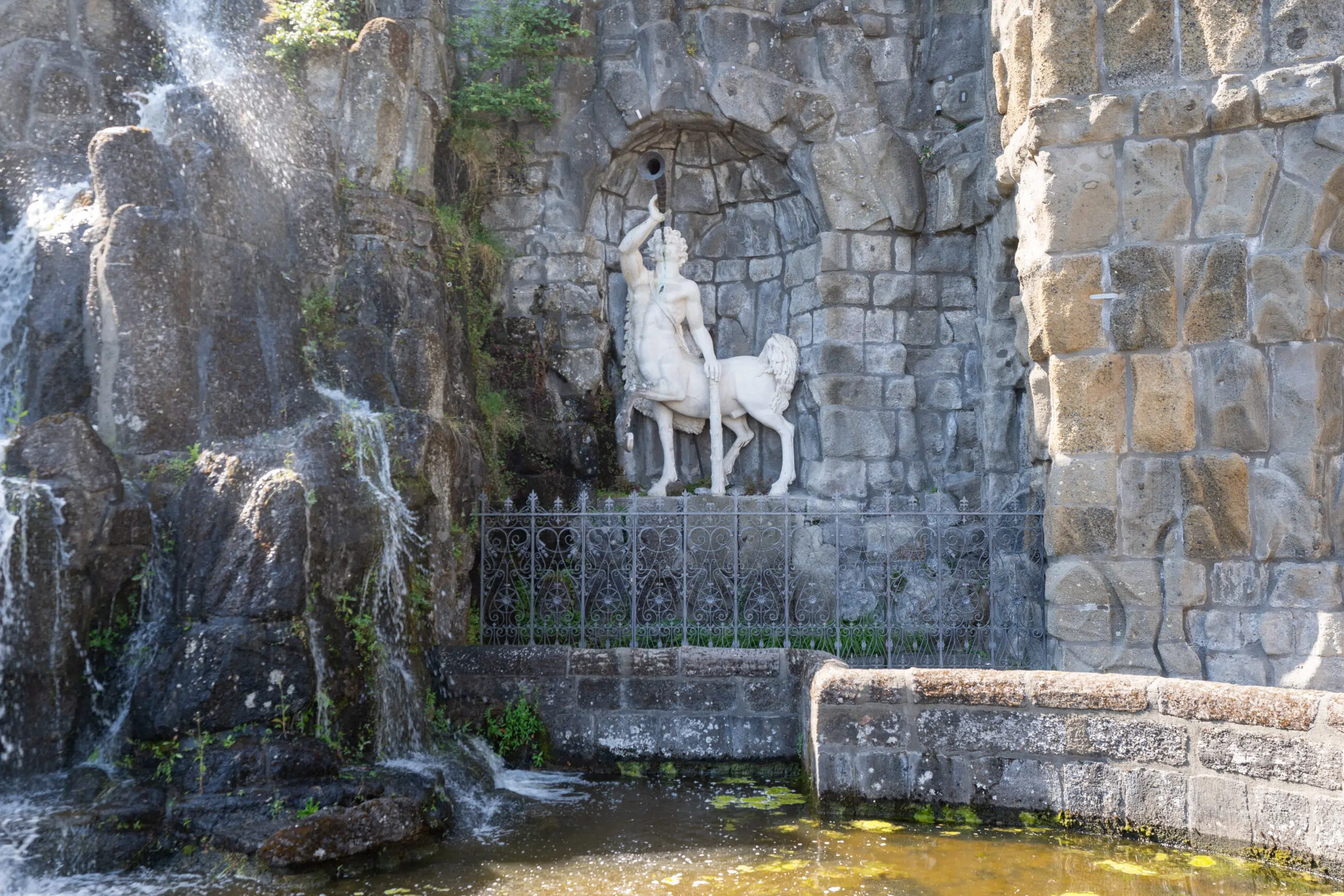A stone sculpture of a centaur holding a horn is set within a rocky alcove, surrounded by cascading water and ivy. The scene feels serene and mythical.
