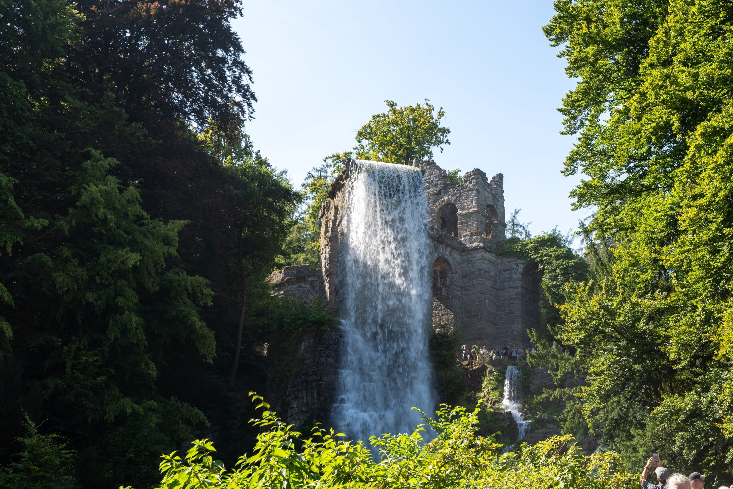 A tall waterfall cascades over a rustic stone structure surrounded by lush greenery under a clear blue sky, creating a tranquil, scenic view.