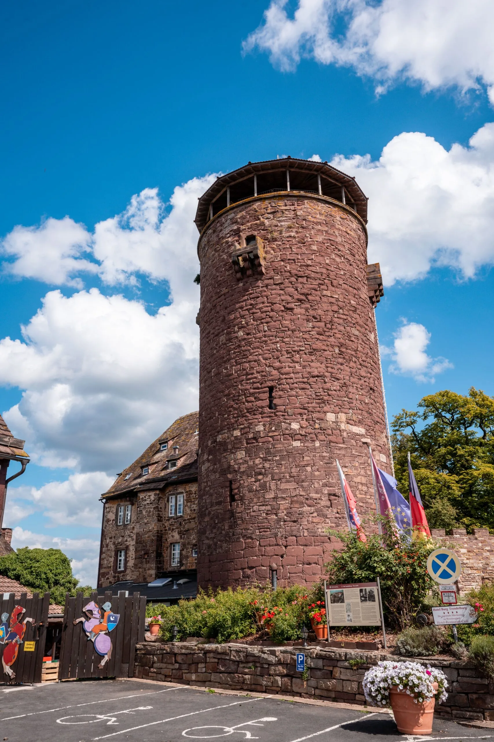 A tall, cylindrical red brick tower with a conical top stands under a bright blue sky with fluffy clouds. In the foreground, colorful flowers and flags adorn the entrance, creating a vibrant and historical atmosphere.