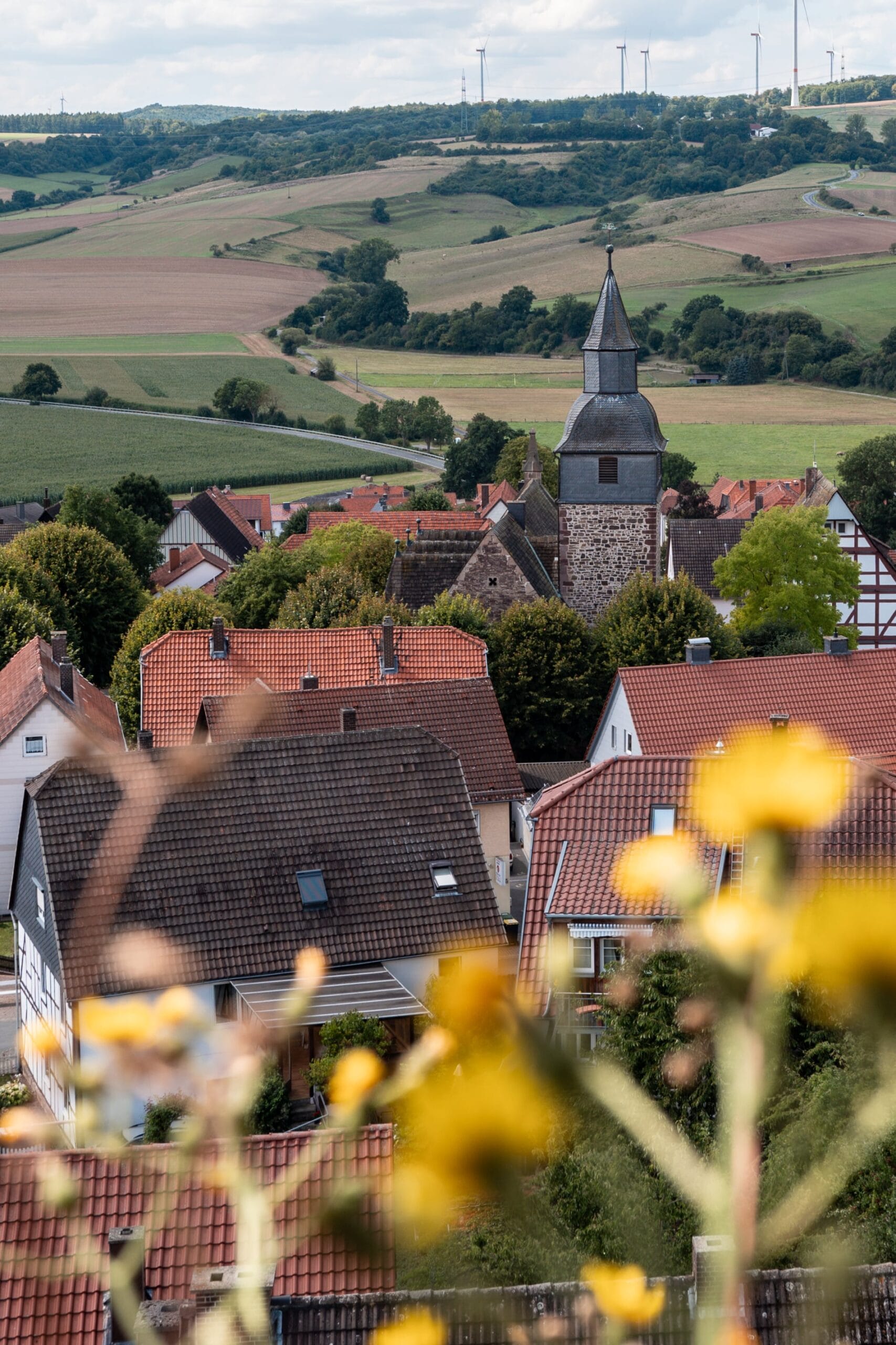 Scenic view of a quaint village with red-roofed houses and a stone church steeple, set against rolling green hills and distant wind turbines. Warm, serene atmosphere.