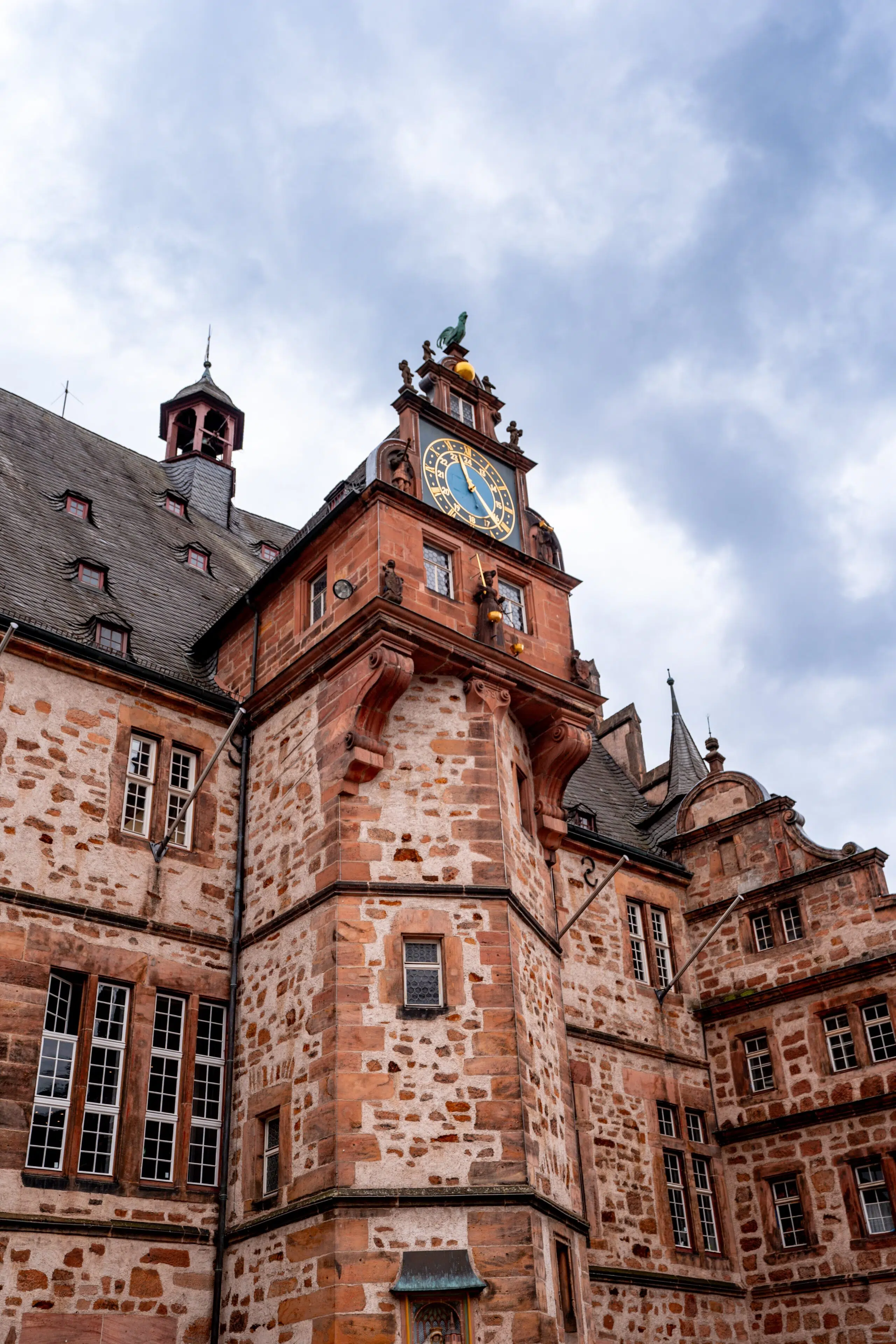 Historic stone building with a clock tower under a cloudy sky. The architecture features red brickwork and steep roofs, creating a timeless and grandiose feel.
