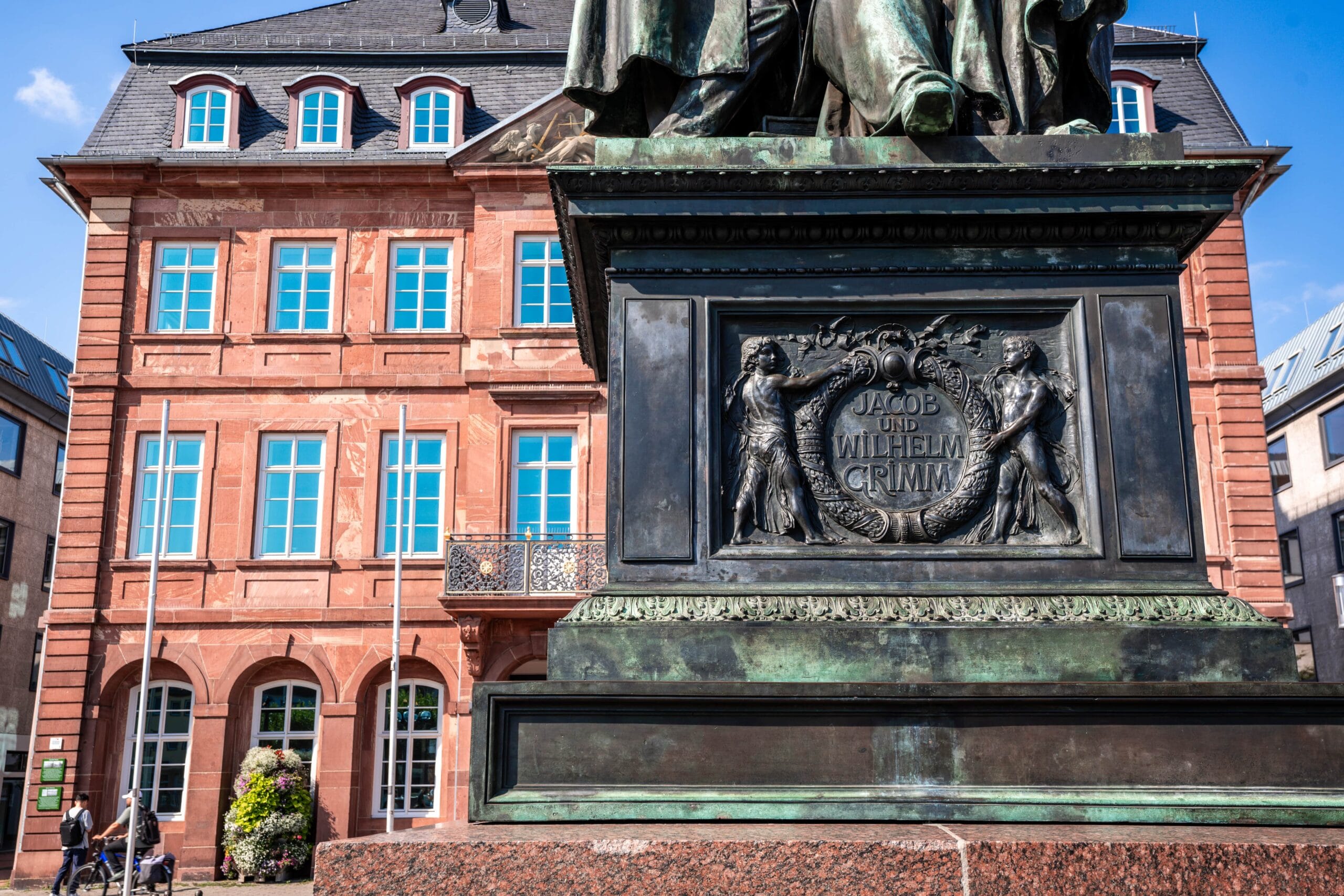 Bronze statue base with "Jacob und Wilhelm Grimm" inscription, framed by figures, in front of a red-brick building with blue windows. Bright, sunny day.