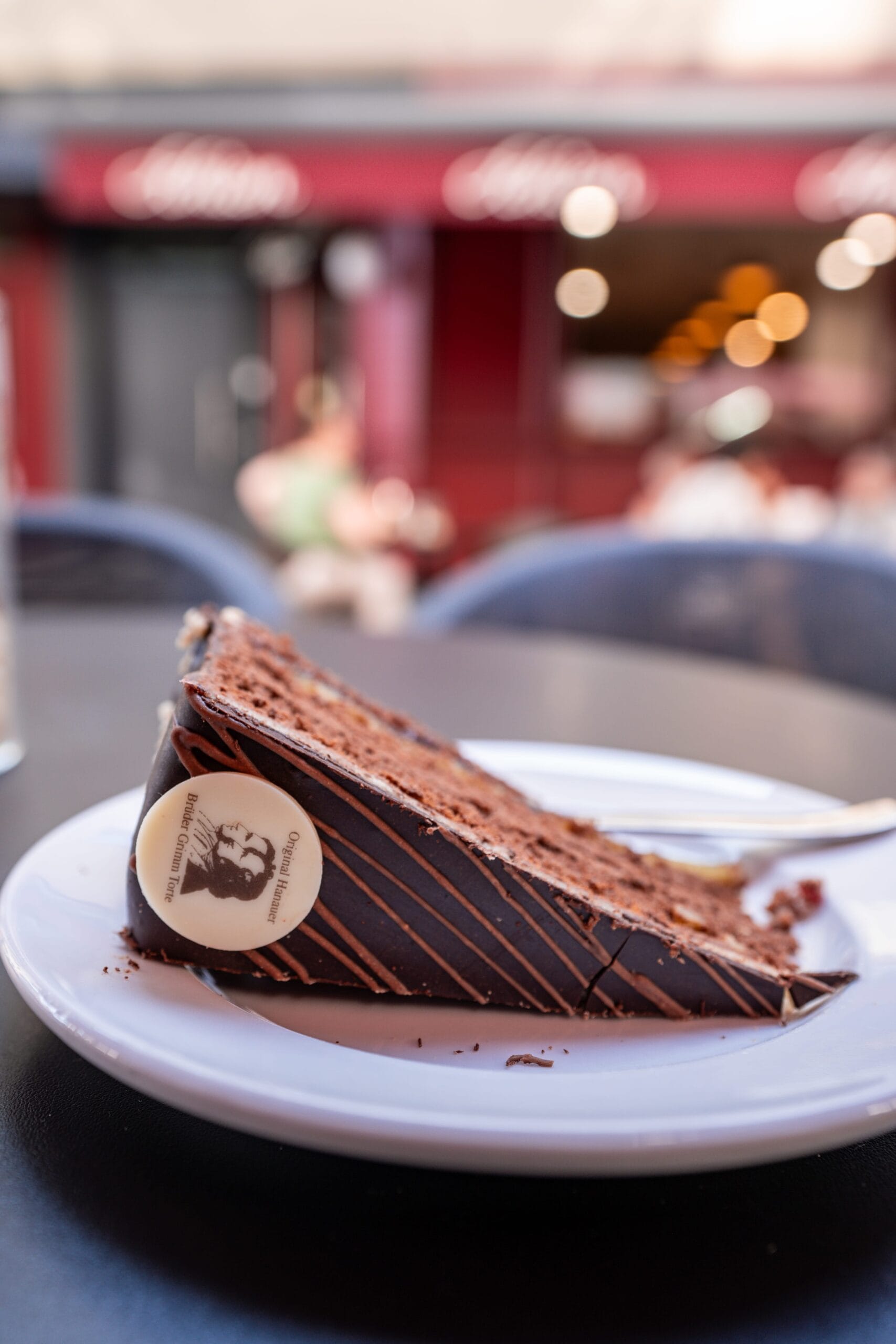 A slice of rich chocolate cake on a white plate with a café backdrop. The cake has a glossy glaze and a chocolate medallion, conveying indulgence.