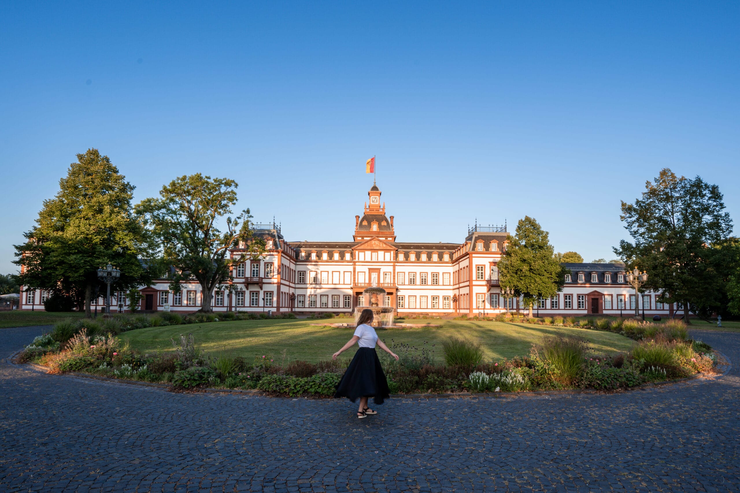 A woman in a black skirt walks toward a grand, historic building with a central tower and flag. It's surrounded by a garden and trees under a clear blue sky.