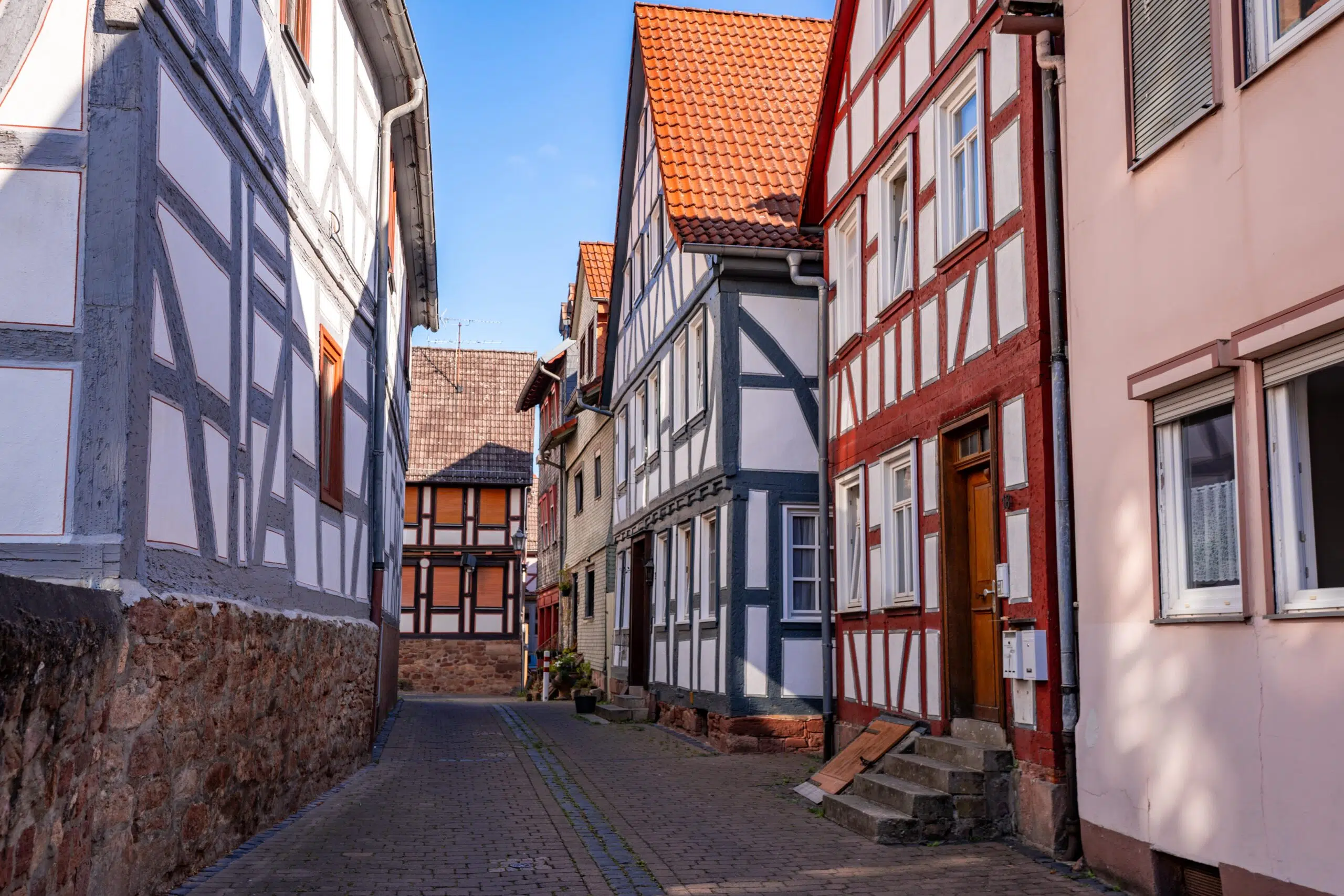 Narrow cobblestone street lined with traditional half-timbered houses featuring red and gray beams, under a clear blue sky, creating a quaint, serene vibe.
