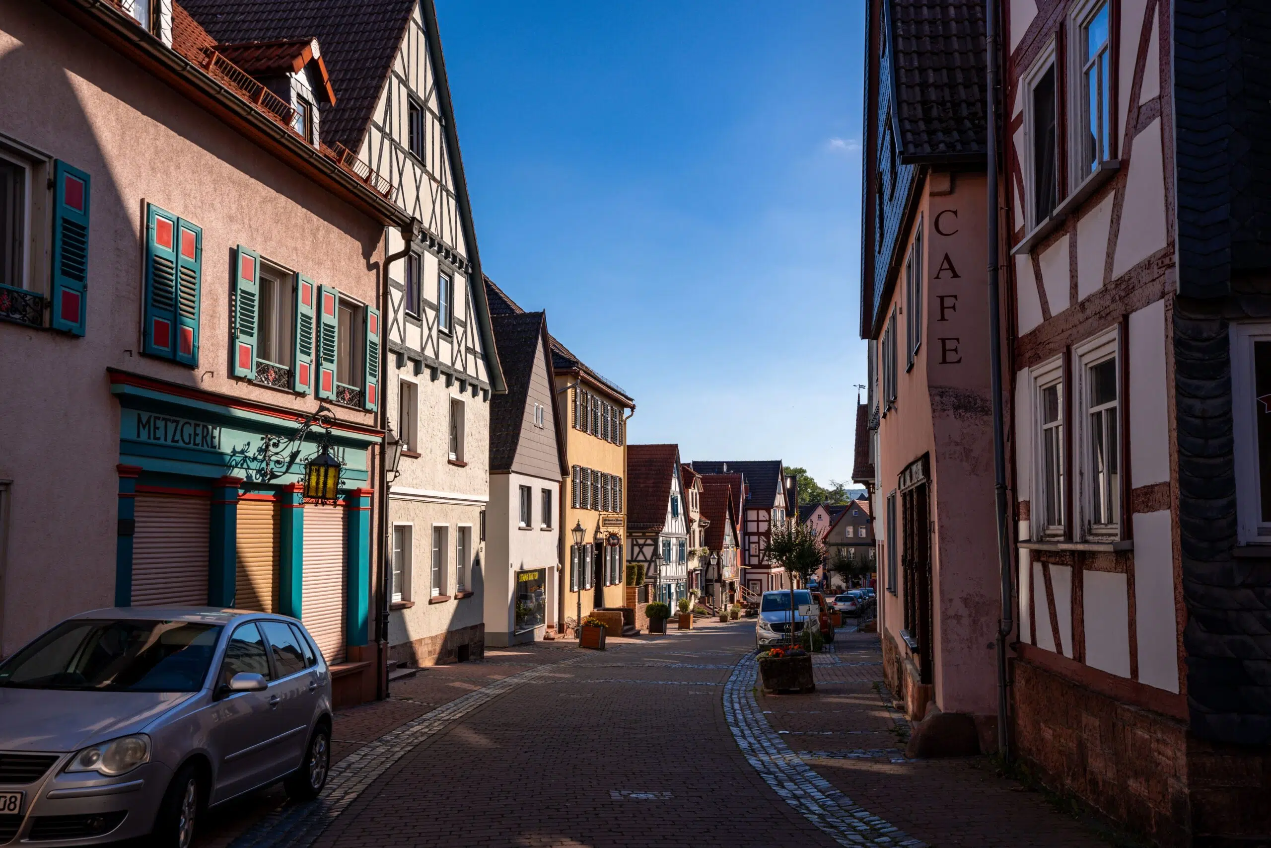 A quaint European village street bathed in sunlight, lined with colorful half-timbered buildings, including a café and a butcher shop, under a clear blue sky.